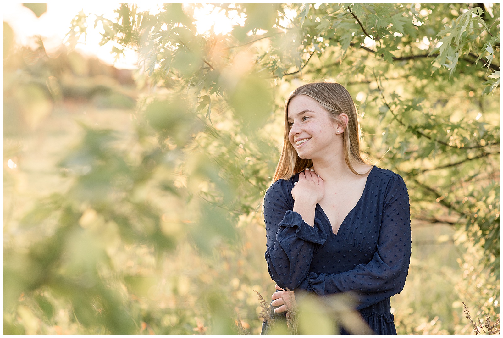 senior girl wear navy blue dress surrounded by trees with sun setting behind her as she looks right holding right shoulder with right hand