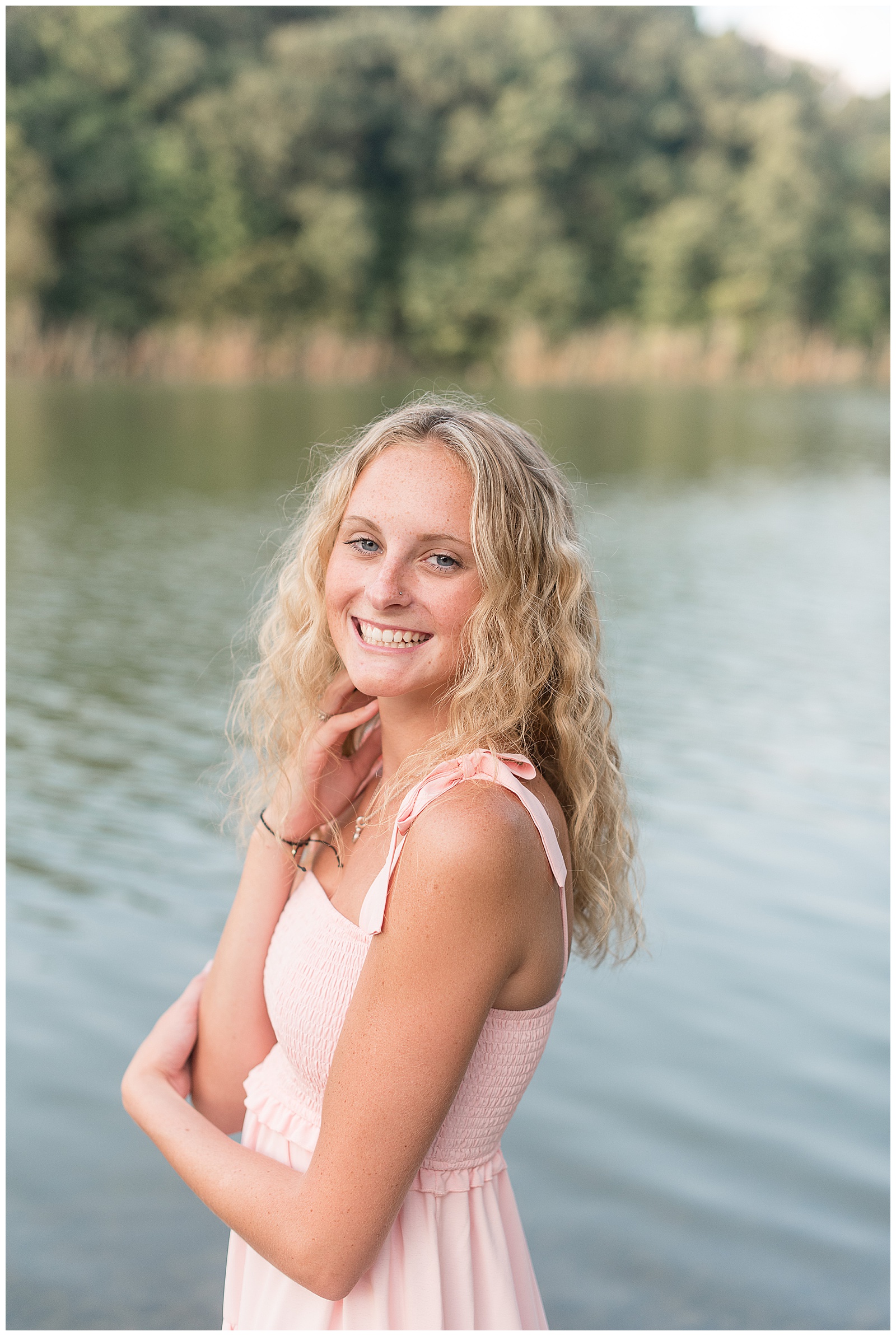 senior girl with left shoulder towards camera and her right hand on her right shoulder smiling wearing light pink dress by lake in lititz pennsylvania