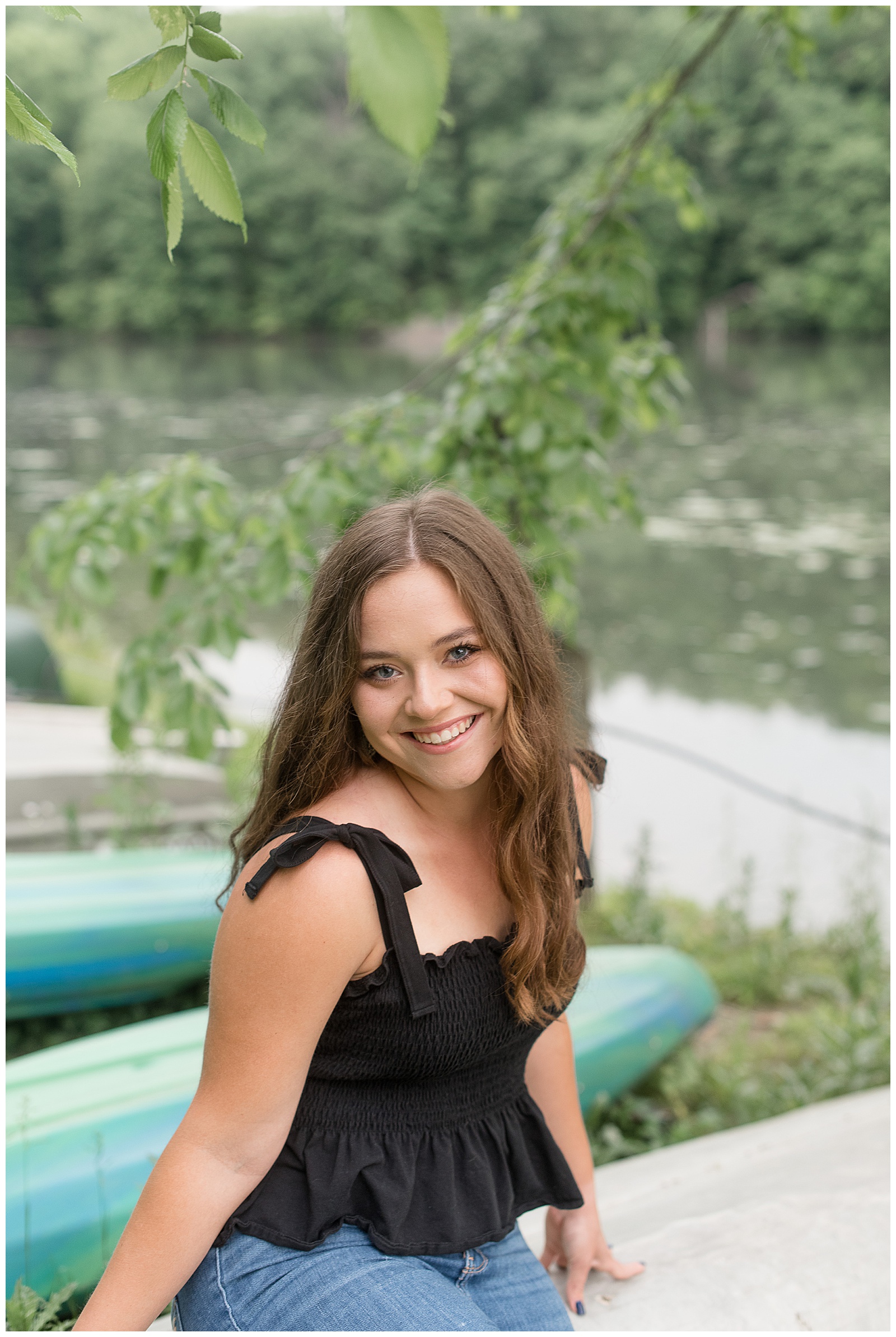 senior girl sitting by row of colorful canoes wearing black tank top and jeans smiling at camera at speedwell forge lake