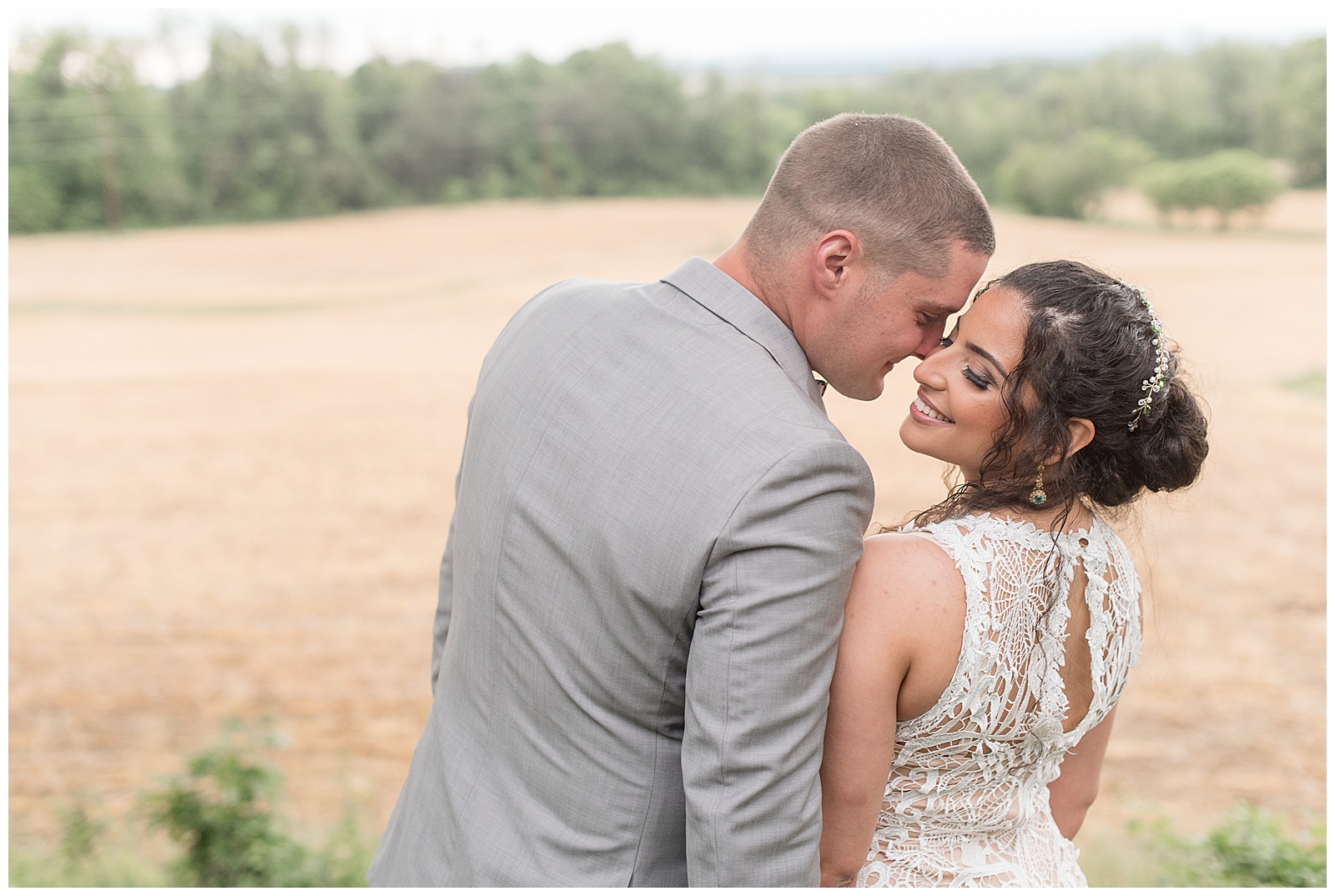 backs of groom and bride toward camera as he leans in to kiss her on her right cheek as she looks back as they stand by field