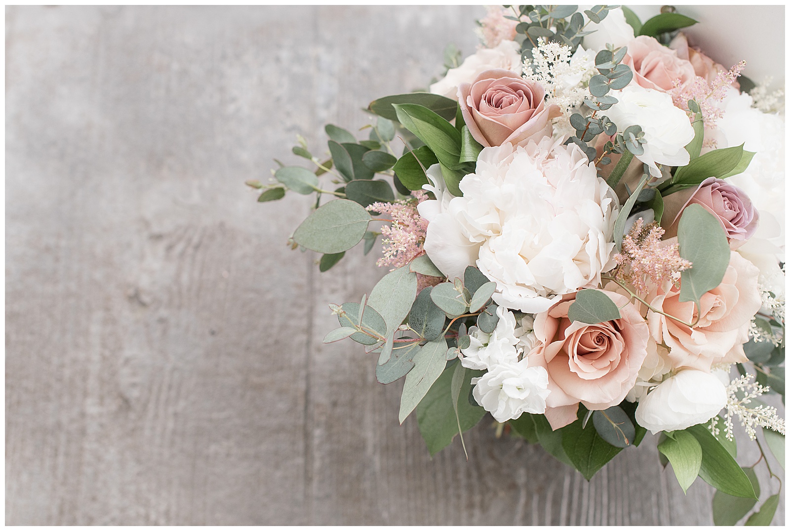 overhead photo of bridal bouquet sitting on gray wooden floor filled with white peonies, light pink roses, and eucalyptus