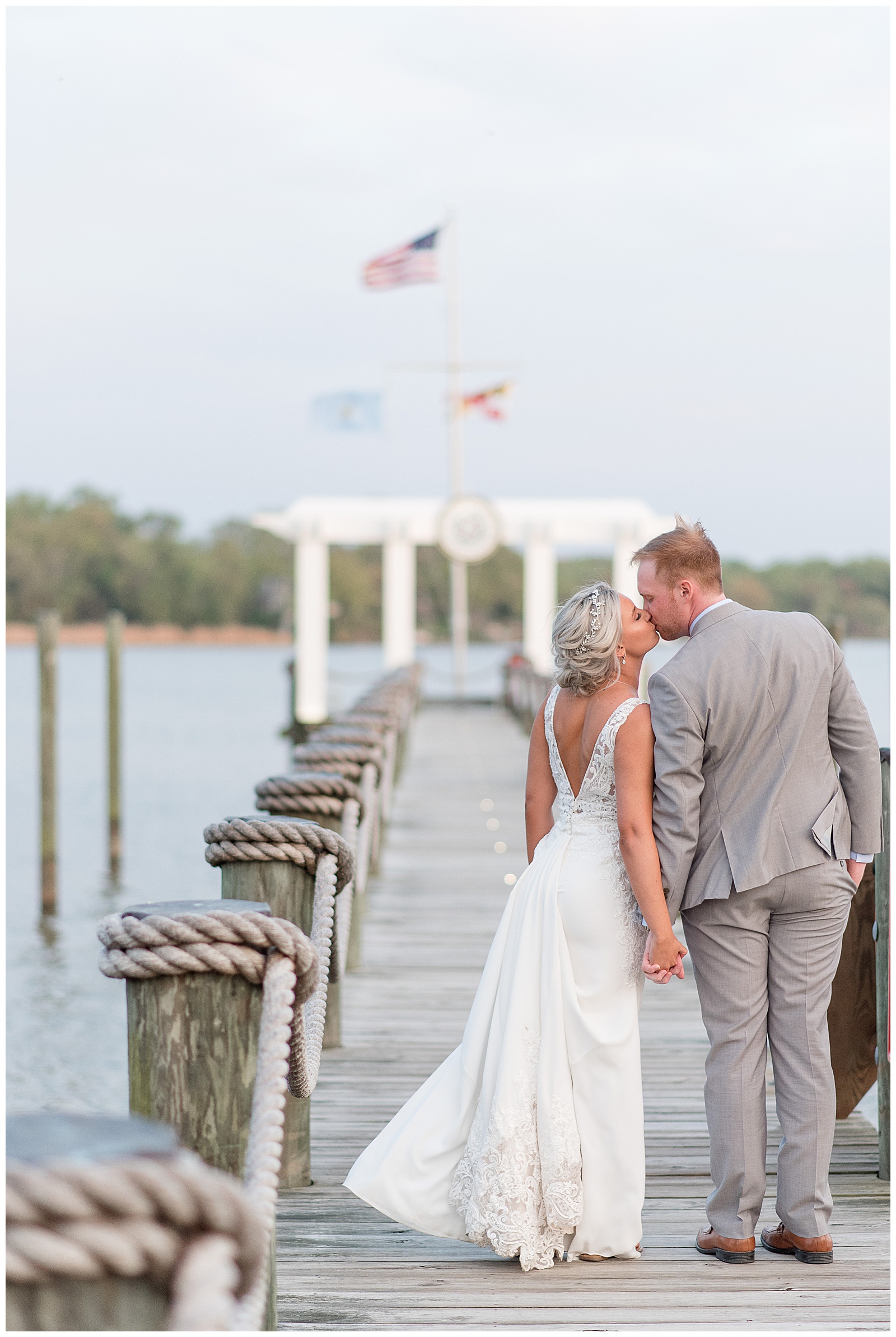bride and groom kissing on wooden dock with their backs toward the camera by beautiful bay on sunny summer day