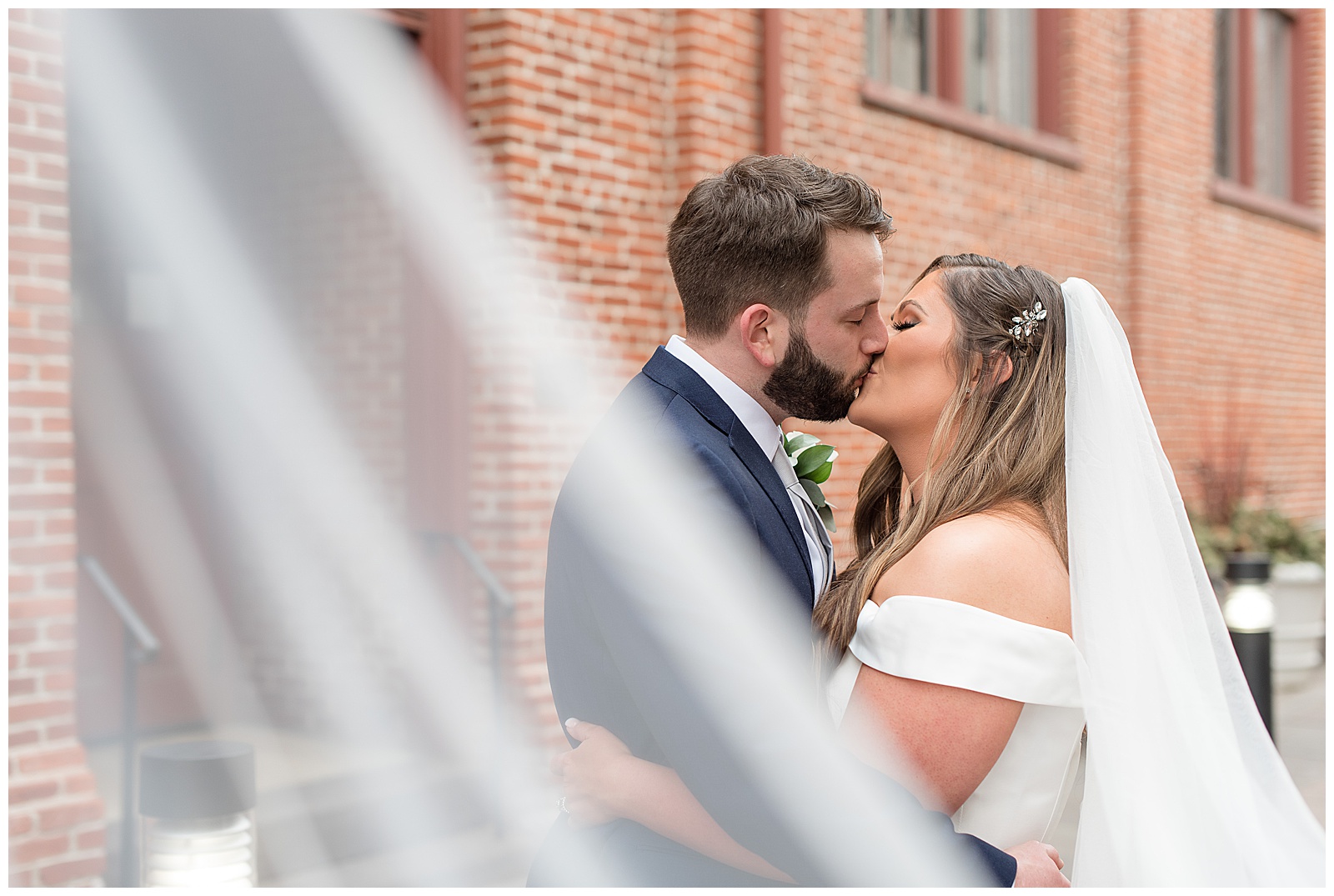 groom and bride kiss in downtown lancaster city with her long bridal veil blowing around them and in front of camera
