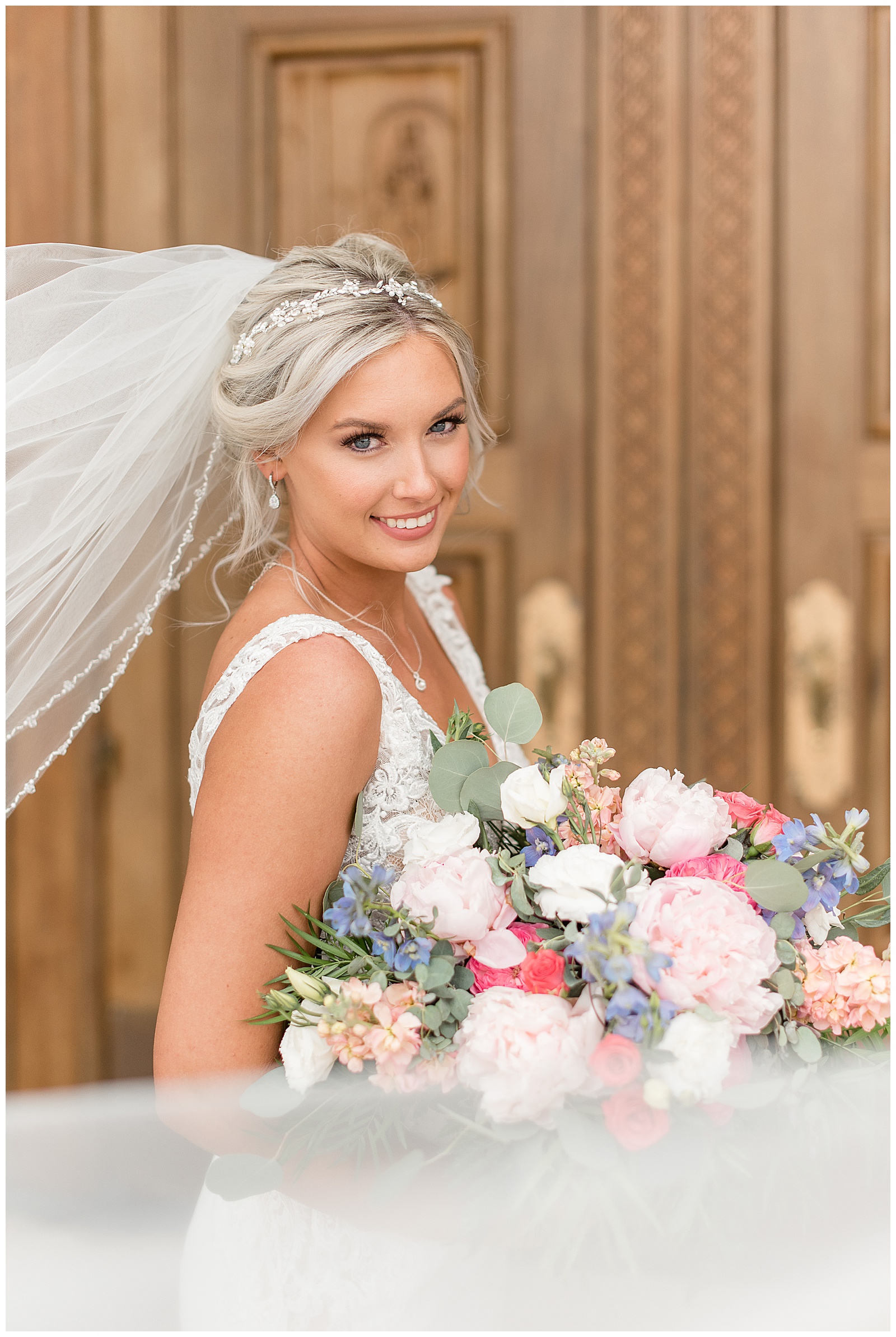 bride smiling at the camera with veil blowing behind her and stunning colorful bouquet displayed in front of her by wooden door