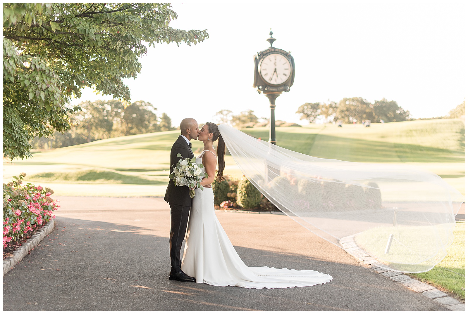 groom and bride kissing by unique clock at the north jersey country club with bride's long veil blowing behind her
