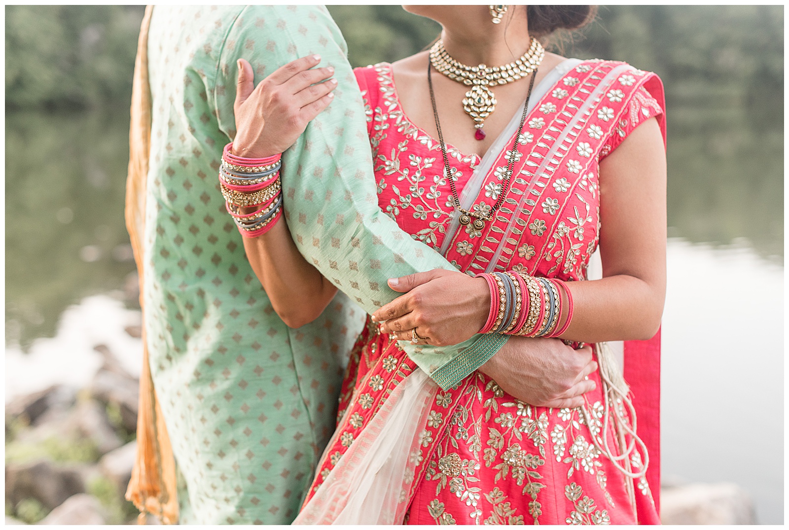 close up photo of groom and bride wearing traditional brightly colored indian wedding attire with their arms wrapped around one another with pond behind them