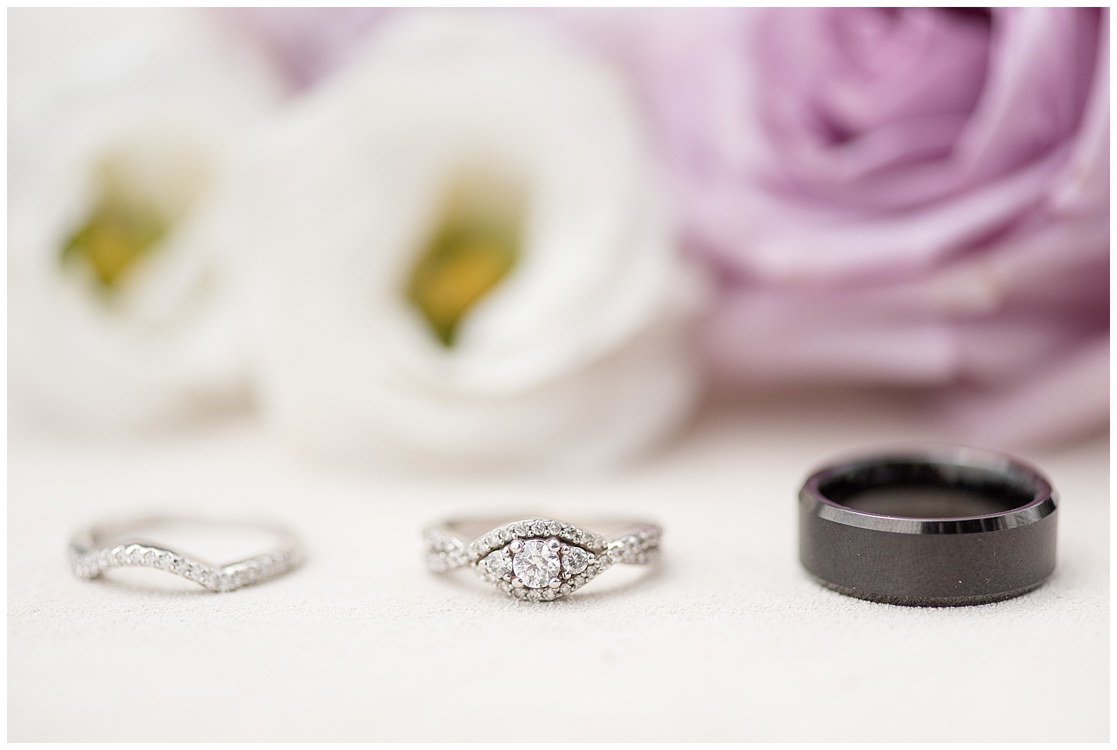 diamond engagement ring in the center with diamond wedding ring to the left and groom's black wedding band to the right with white and purple flowers in background