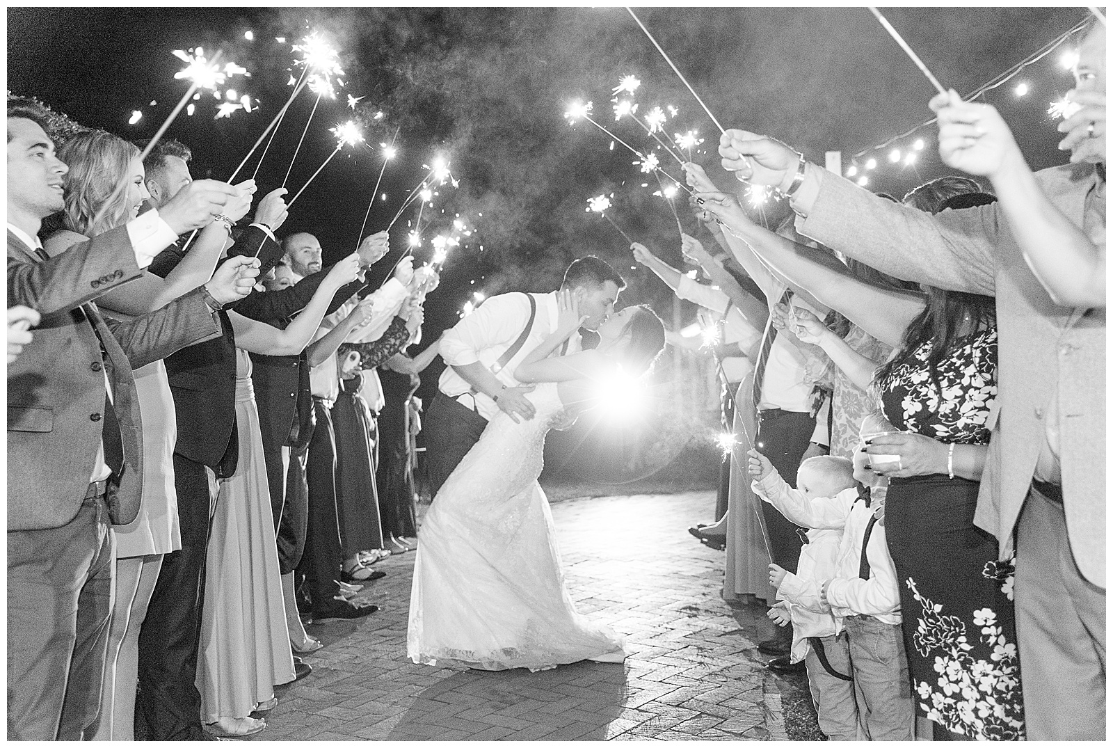 black and white photo of groom dipping back his bride as they kiss in between two rows of guests holding up long sparklers above them