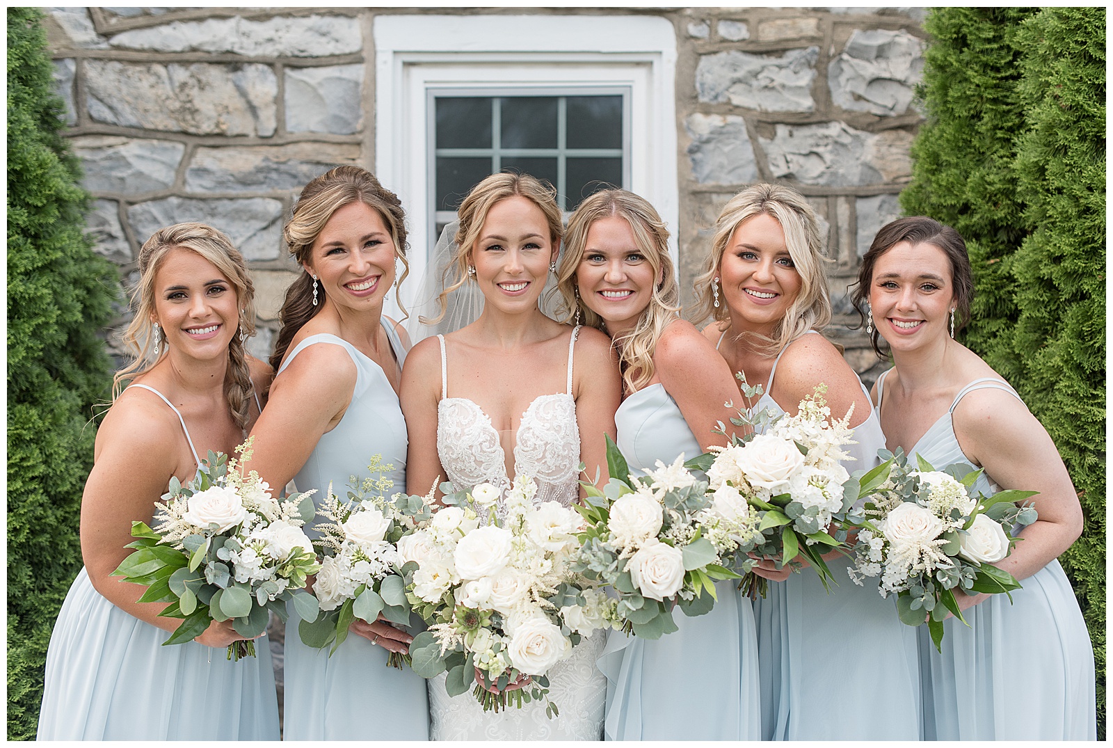 bride surrounded by her five bridesmaids wearing light blue gowns and everyone holding bouquets by historic stone building