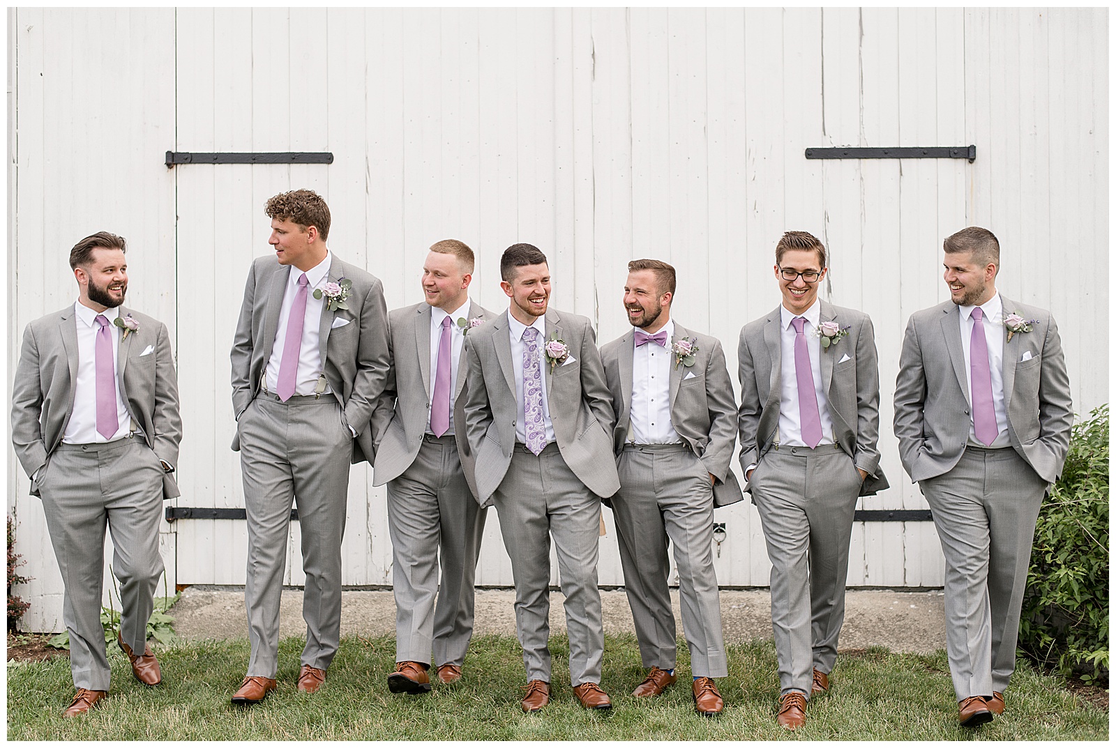 groom with his groomsmen all wearing light gray suits with purple ties and walking toward camera with hands in pockets and looking in different directions by white barn