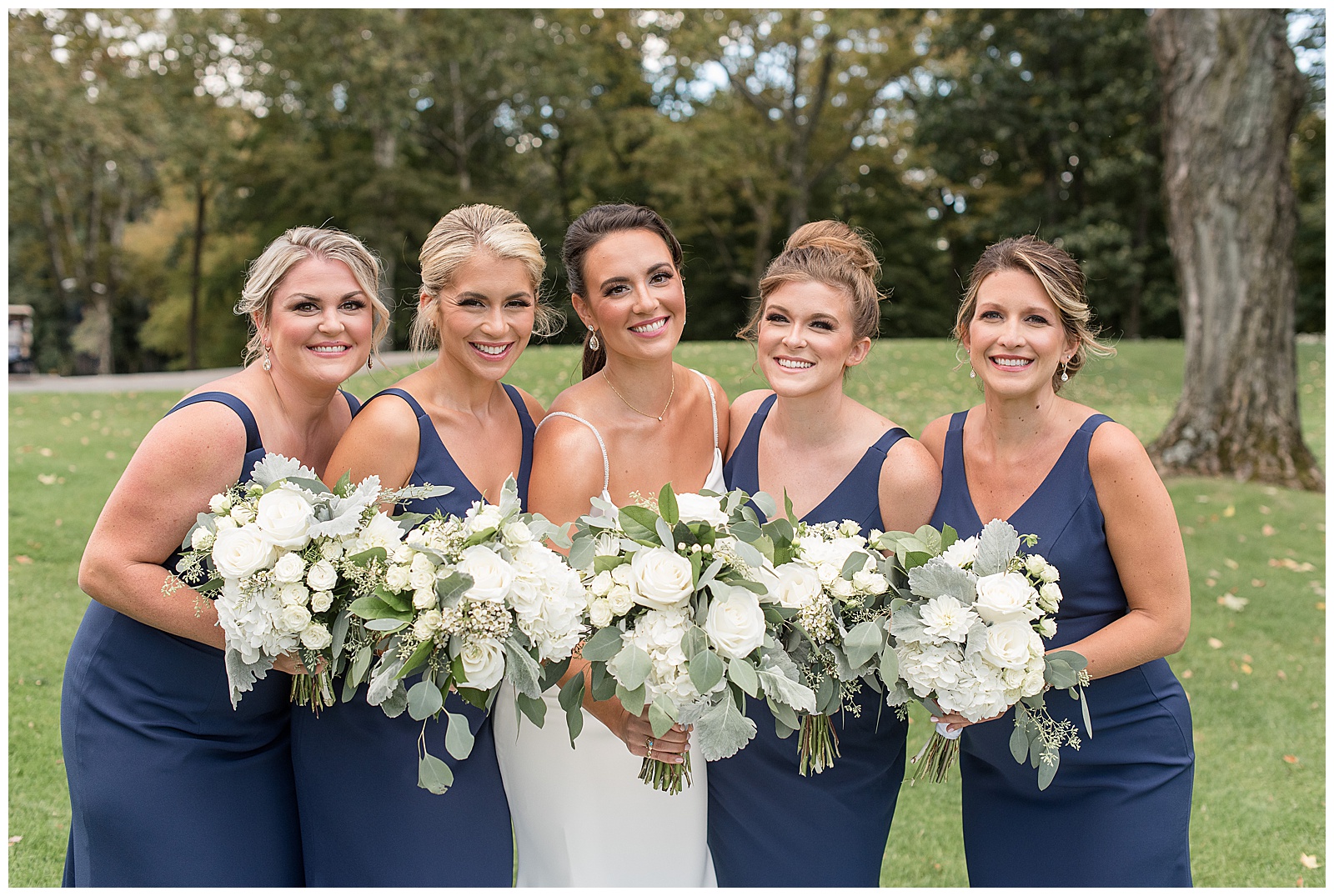 bride in white sleeveless gown surrounded by bridesmaids in dark teal sleeveless gowns all leaning in holding white bouquets