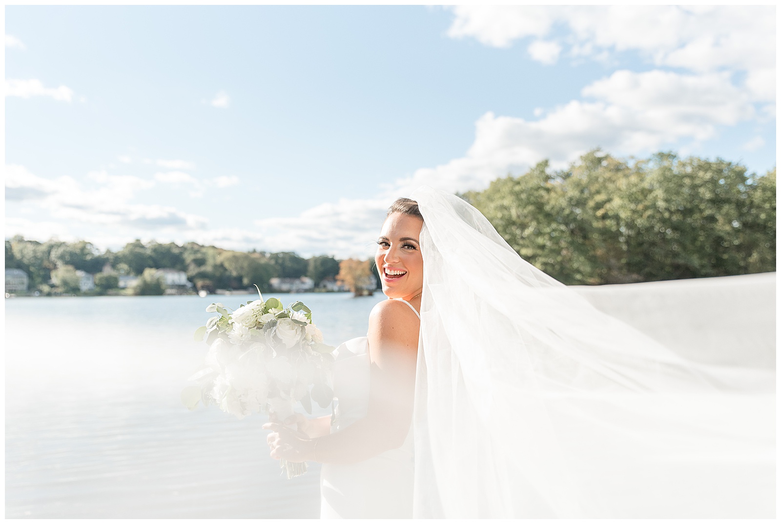 bride standing on dock by water on bright sunny day with long veil blow all around her in new jersey