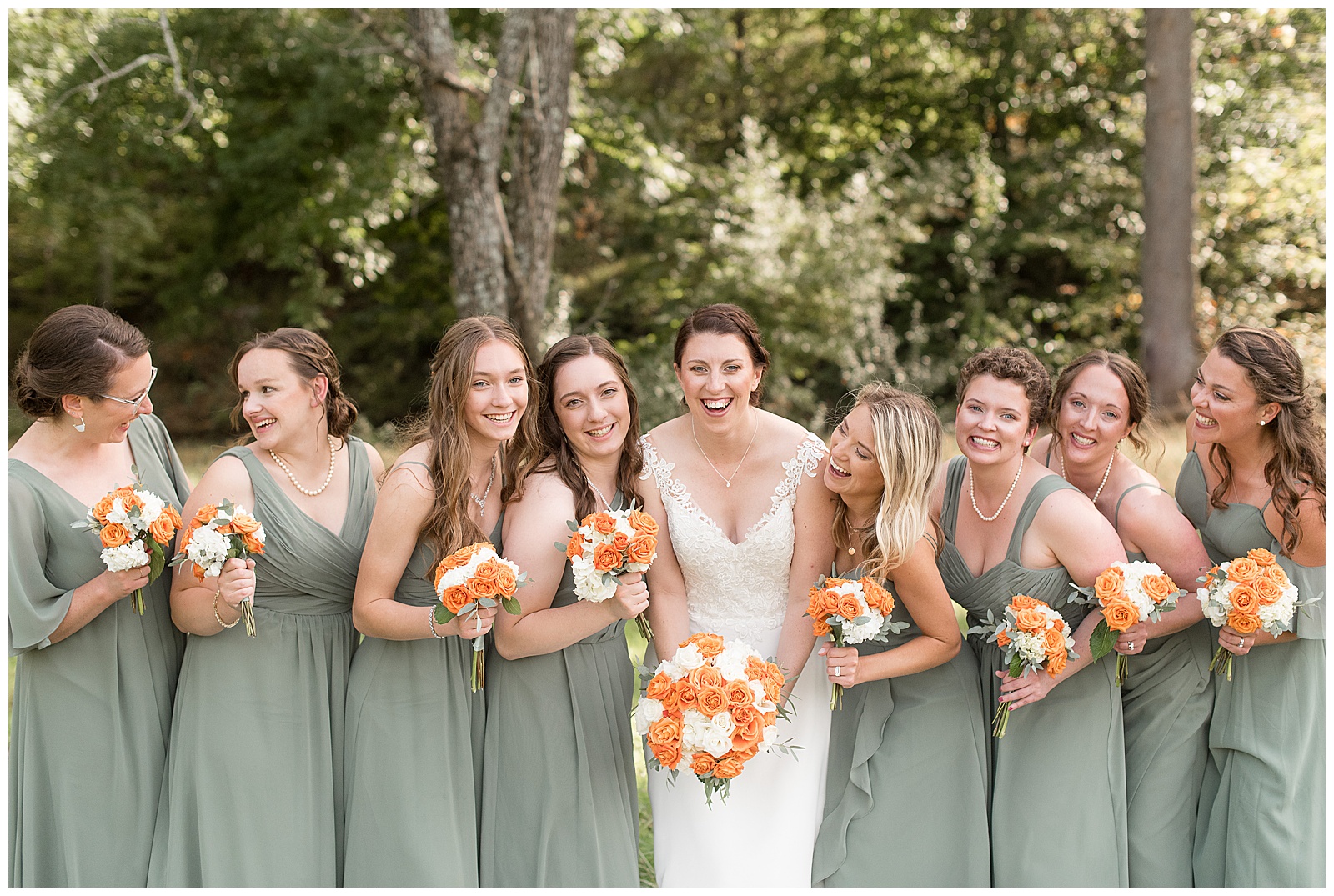 bride in white wedding gown surrounded by bridesmaids wearing light green dresses all lean in and smile at camera as they hold orange and white bouquets
