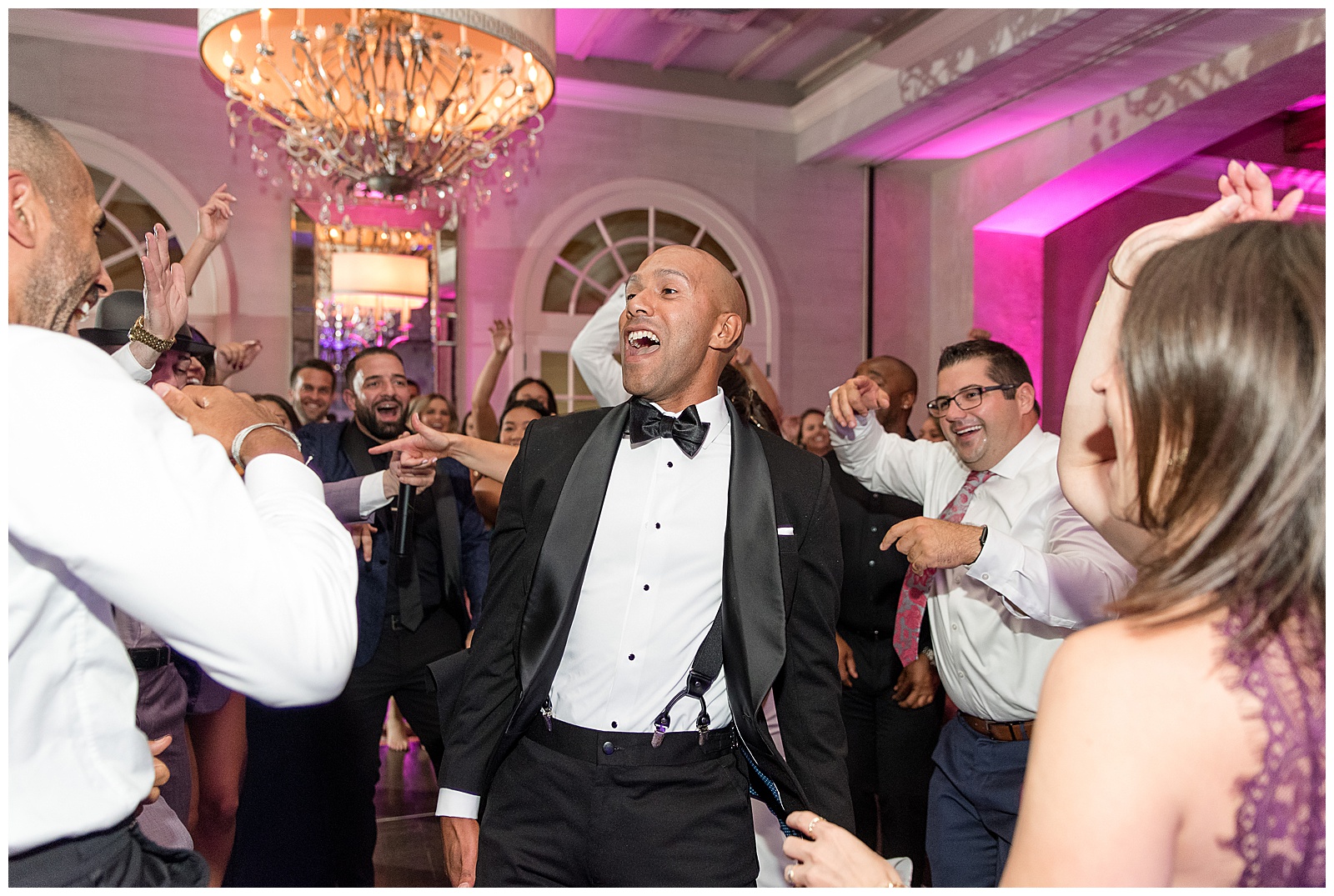 groom dancing and smiling during wedding reception surrounded by guests with large chandelier and pink lighting in the background