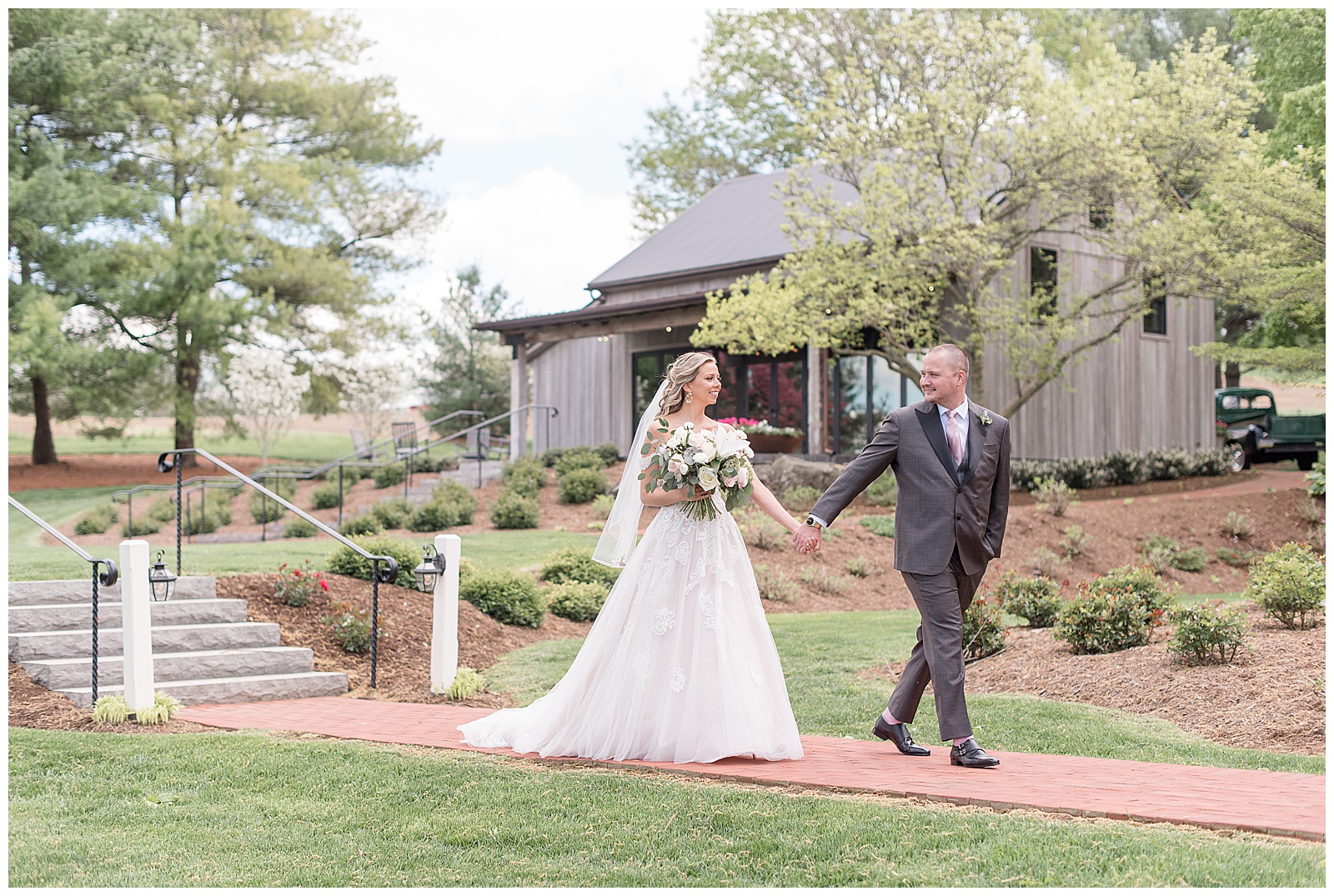 groom leading his bride along brick pathway as she holds bouquet at beautifully manicured property on sunny day