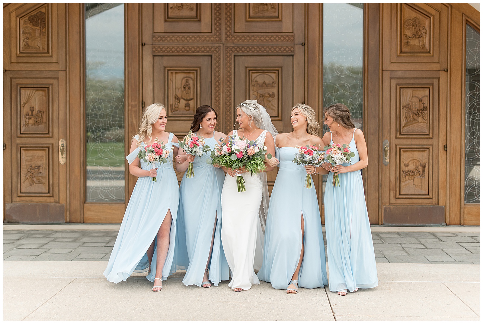 bride with her bridesmaids wearing light blue gowns all walking towards camera and smiling and looking at each other as they hold bouquets outside church doors