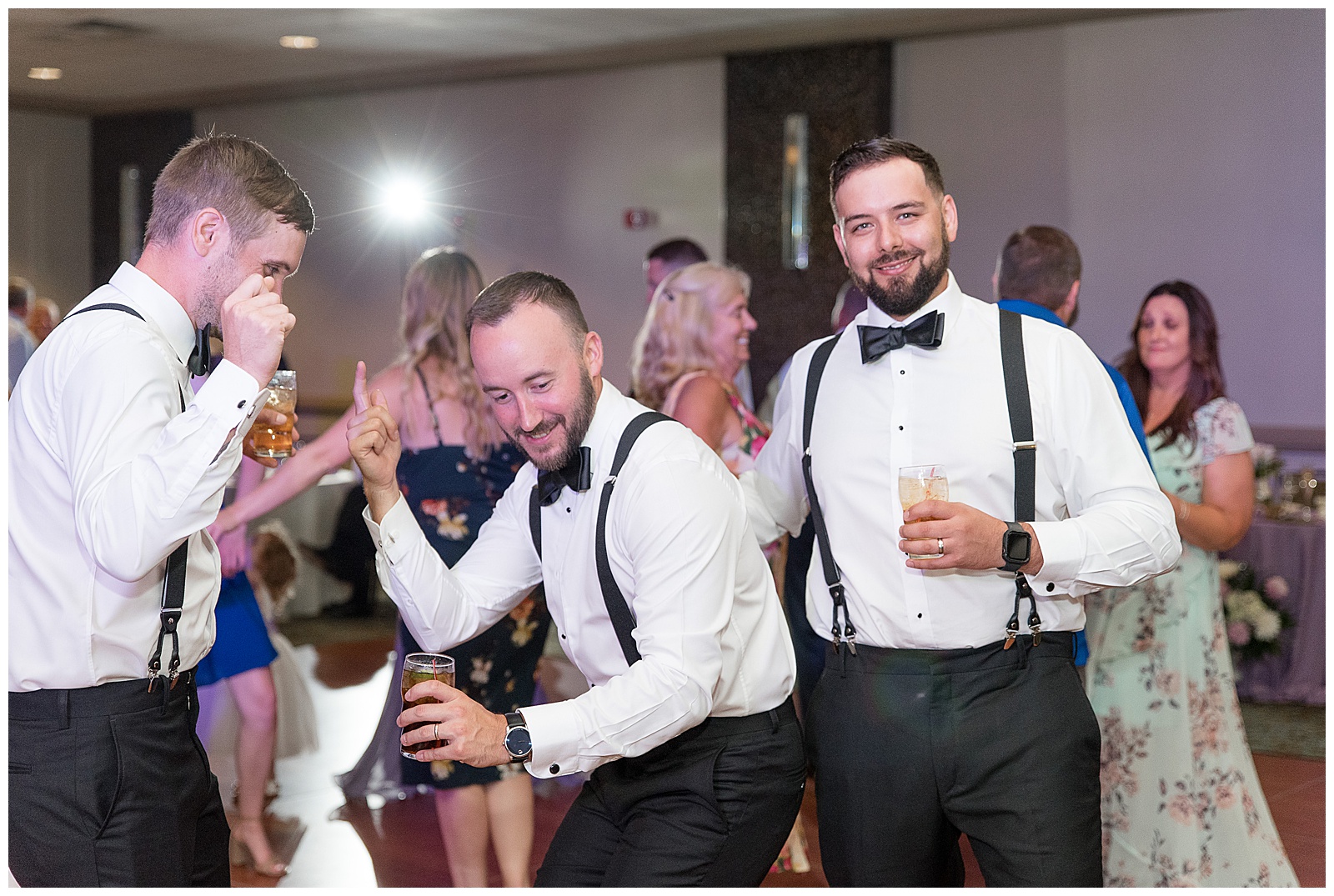 groom and his groomsmen dancing and holding drinks during wedding reception as they all wear white dress shirts and black pants with suspenders