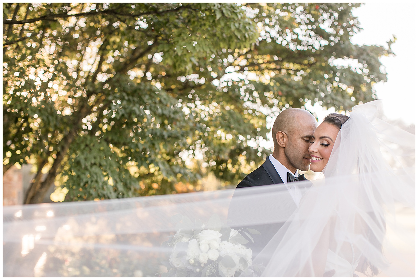 groom kissing bride as she looks over left shoulder and her long white veil swirls around them with trees and sunset in background