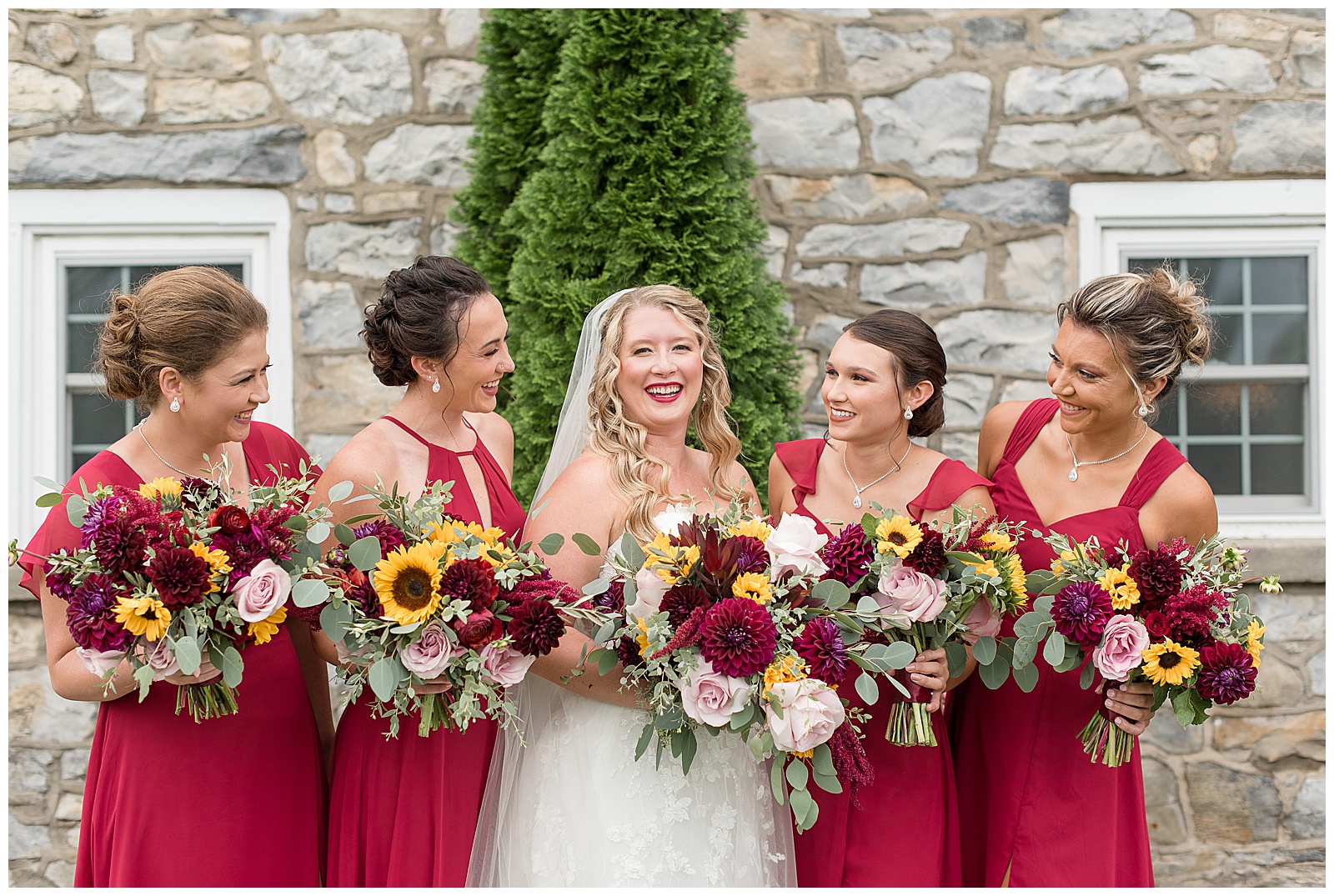 bride smiling at camera as her bridesmaids smile at her in their red dresses with everyone holding flower bouquets