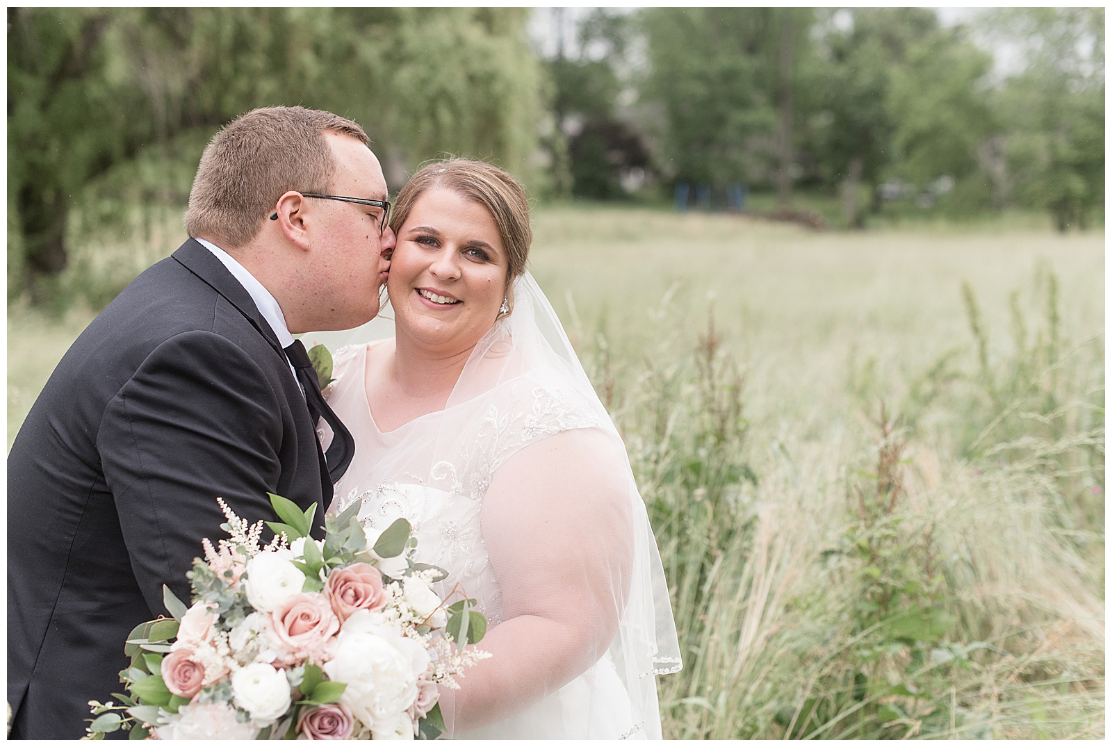 groom kissing his bride on her right cheek as she smiles at camera while they standing in meadow of tall wild grasses at the willows in strasburg pennsylvania