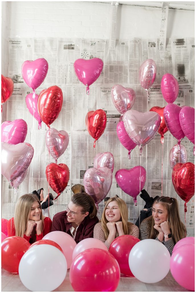 four seniors laying on their stomachs by newspaper-covered wall and surrounded by valentine's balloons at hingeworks