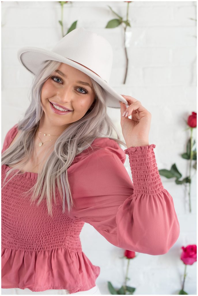 senior girl holding edge of trendy white hat with her left fingers by rose-covered wall at hingeworks in lancaster city