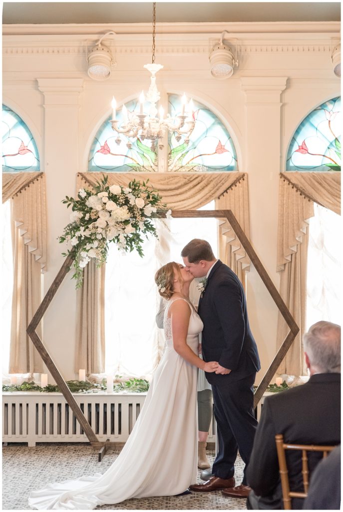 bride and groom sharing their first kiss during wedding ceremony by hexagon floral display inside ballroom at the lititz springs inn and spa