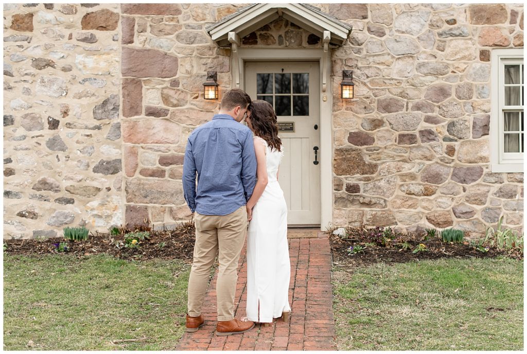 engaged couple standing close with backs toward camera and foreheads touching on brick pathway by stone building at historic poole forge