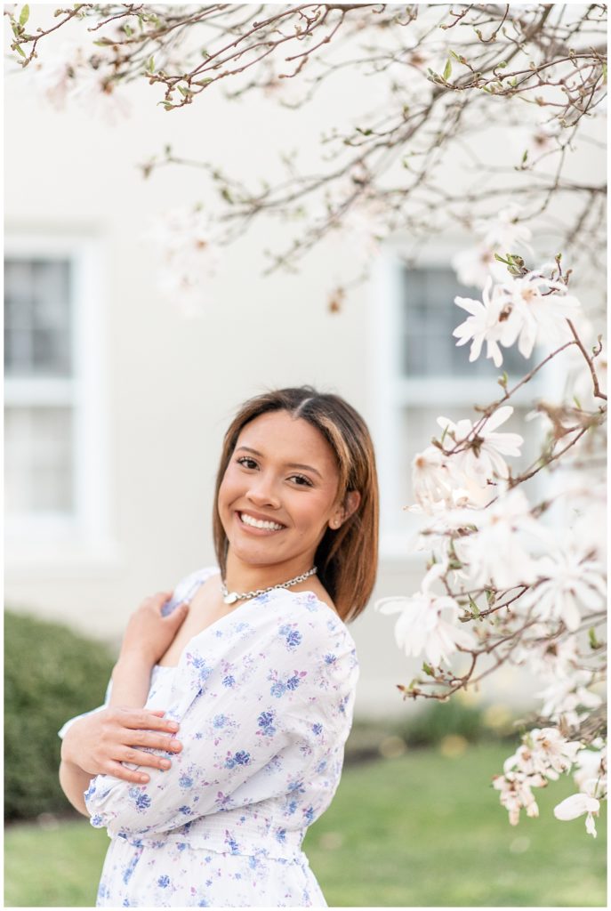 senior girl wearing blue and white floral dress by blooming spring tree in lititz, pennsylvania