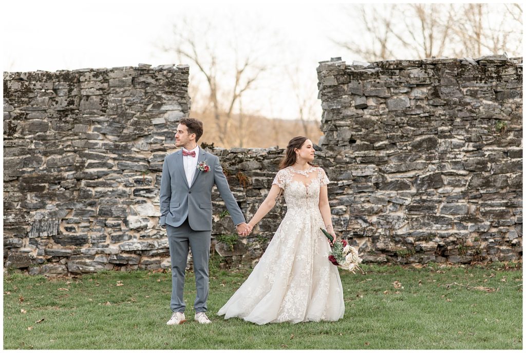 bride and groom holding hands and looking in opposite directions by stone wall in mechanicsburg, pennsylvania