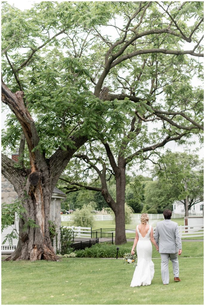 couple holding hands as they walk away from camera by tall trees at the barn at silverstone in lancaster pennsylvania