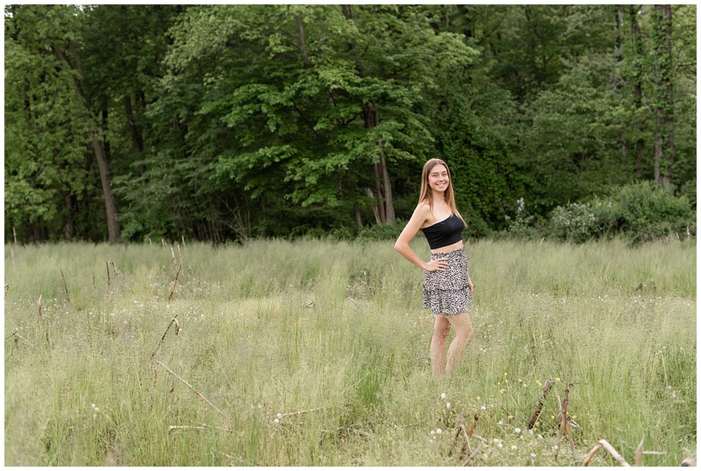 senior girl with right shoulder toward camera and right knee popped with hand on hip smiling in grassy meadow at masonic village