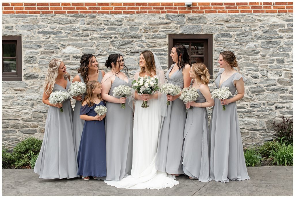 bride surrounded by her bridesmaids and flower girl wearing gray and navy blue dresses by barn