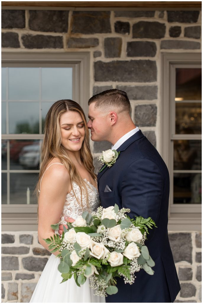 groom going in to kiss bride's left cheek as she looks down and they hug by stone barn wall