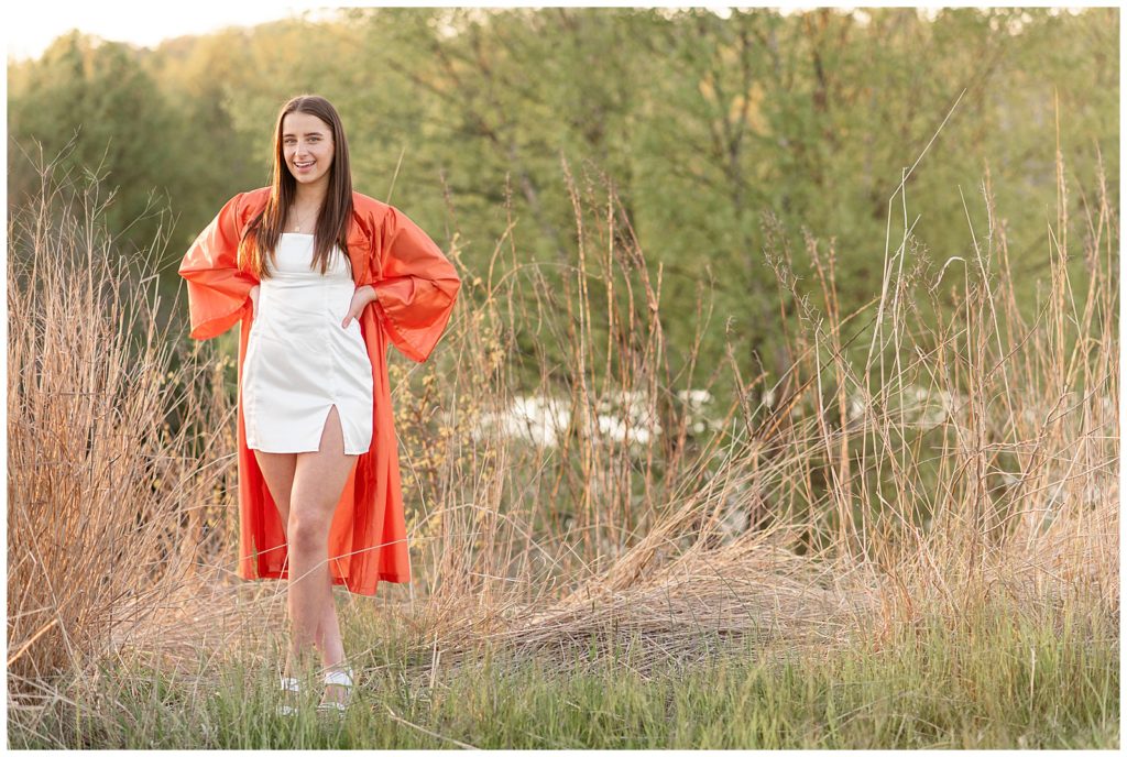 senior girl in white dress with bright orange graduation gown overtop with hands on hips in tall grasses in lancaster county