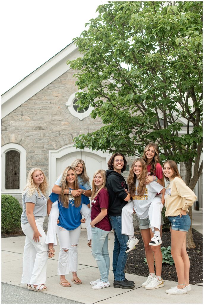 eight class of 2022 seniors standing in opposite directions and some holding one another piggyback style by stone building in lititz pennsylvania