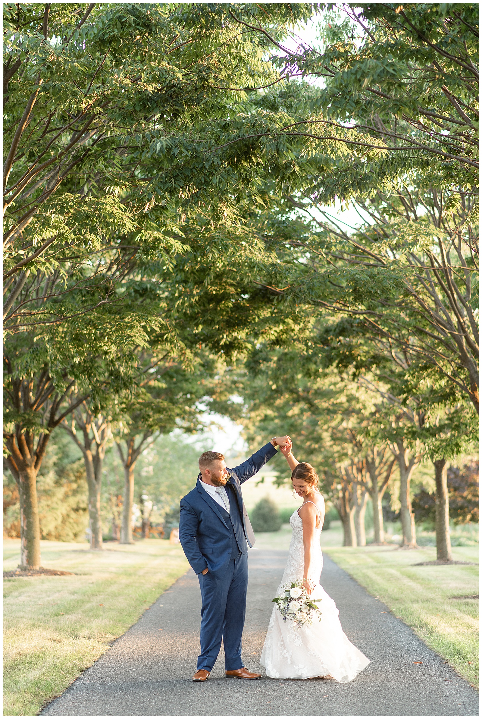 groom twirling his bride under his left hand along tree-lined pathway at lakefield weddings