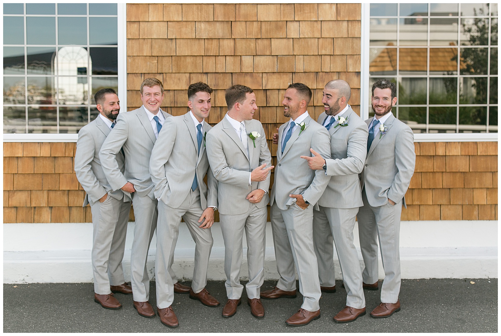 groom and his groomsmen all looking at each other and smiling outside tan building with cedar shake shingles in bay shore, new york