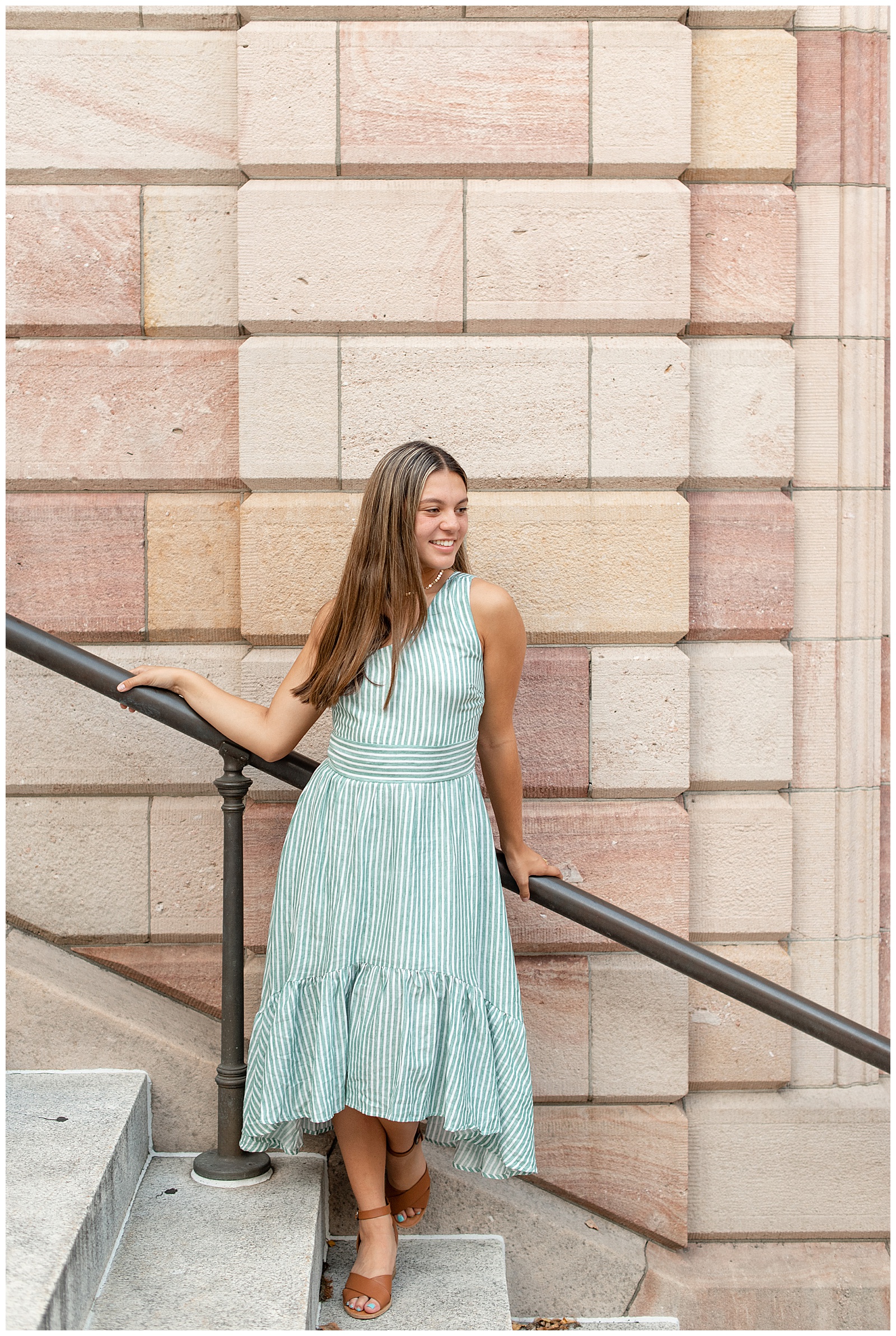senior girl wearing blue one-strap flowy dress holding onto handrail of lancaster county courthouse and looking to her left smiling in downtown lancaster