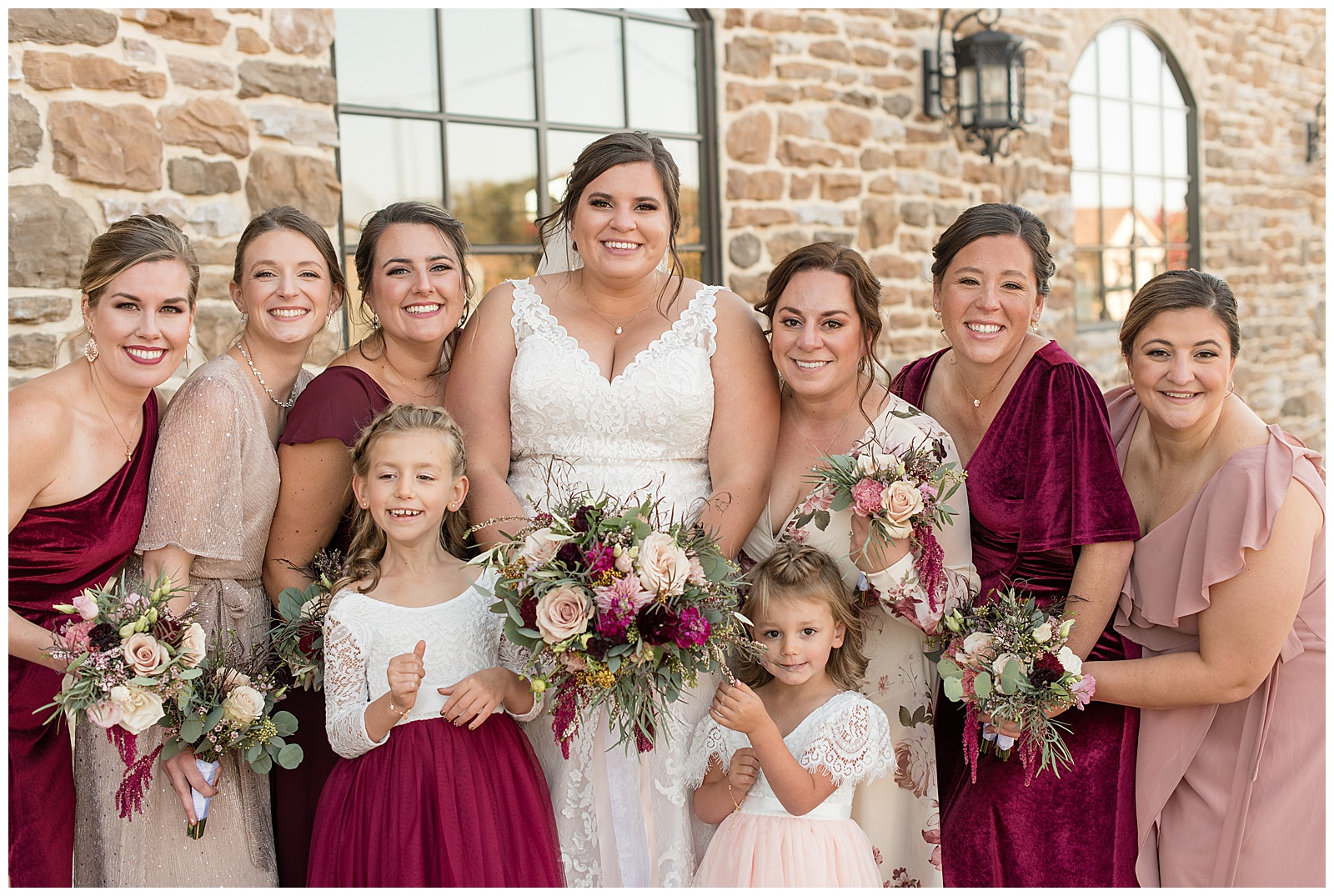bride surrounded by her six bridesmaids in maroon and mauve dresses and two flower girls by stone building in kutztown pennsylvania