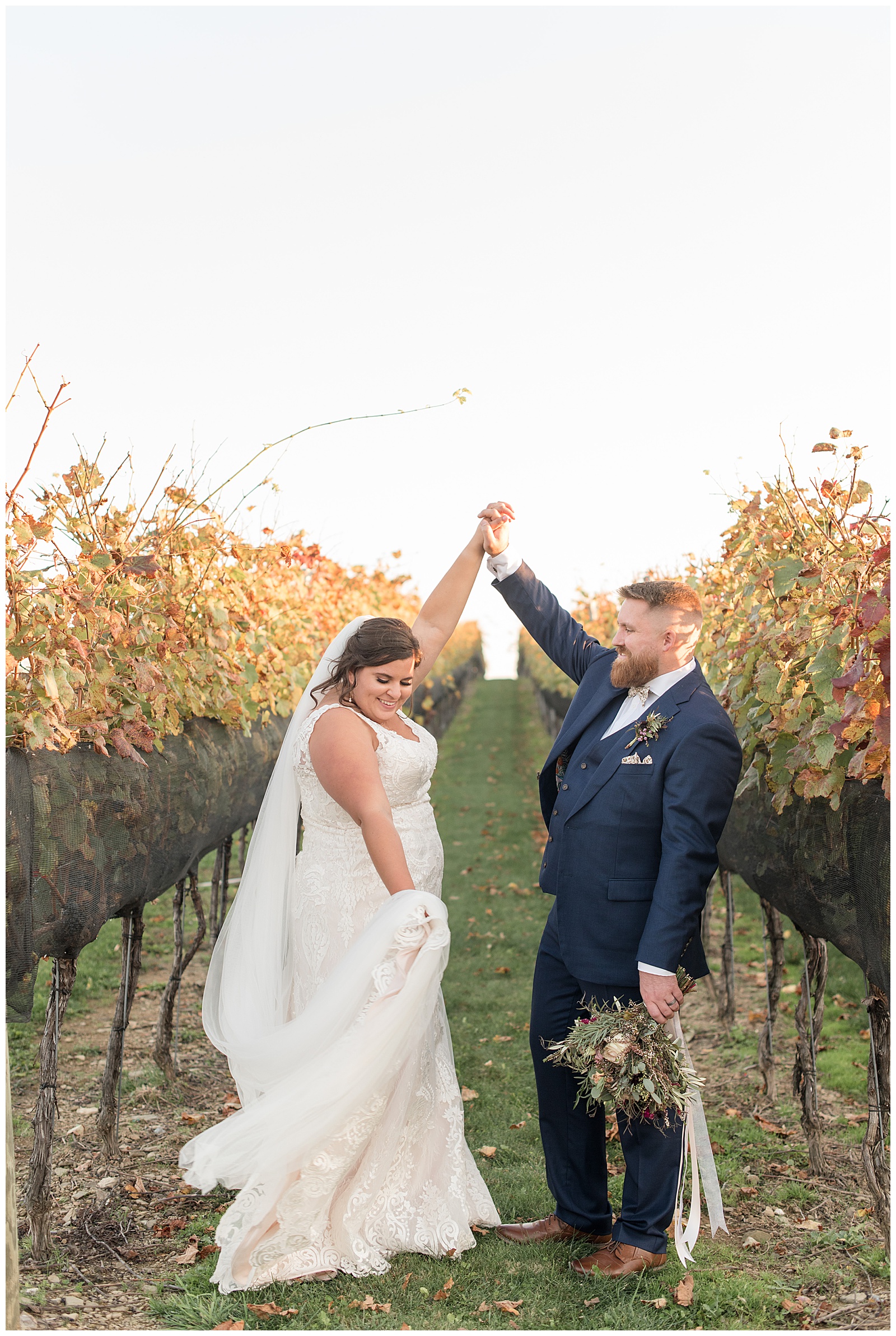 groom twirling his bride under his right hand as she holds her long veil in vineyard row at folino estate