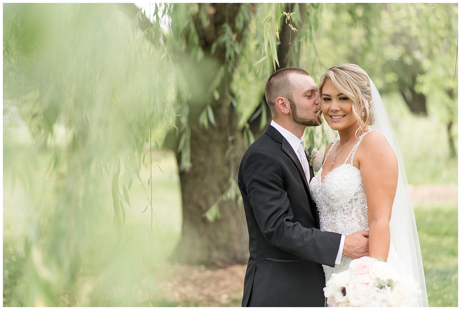 groom kissing his bride's right cheek by willow tree at stoltzfus homestead in lancaster pennsylvania