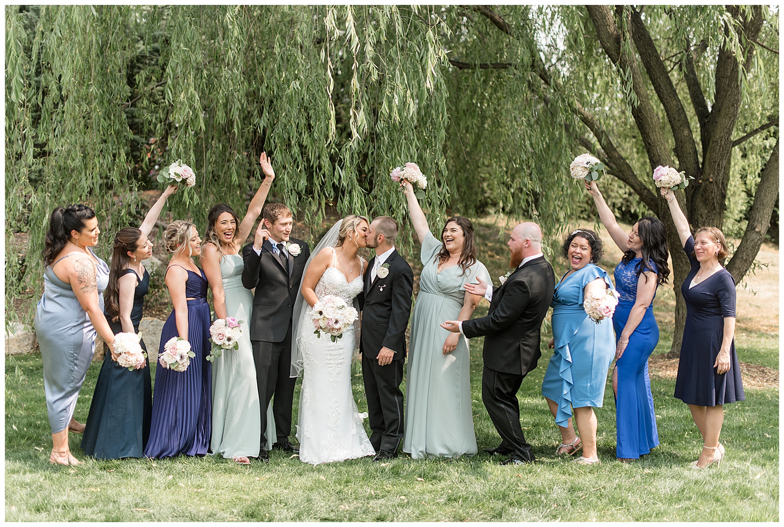 bride and groom kissing surrounded by their cheering bridal party by willow tree at stoltzfus homestead and gardens