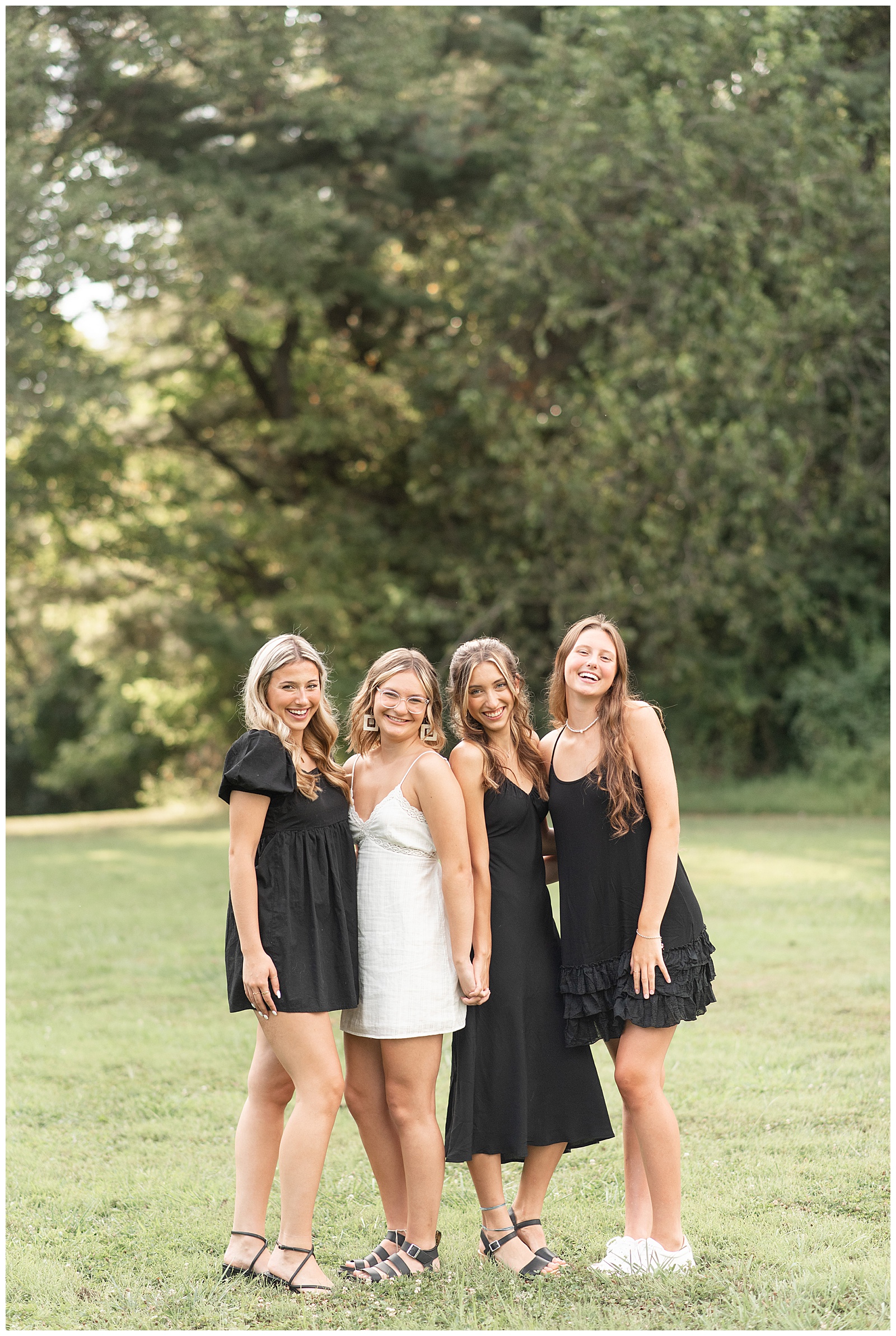 three senior girls in black dresses and one senior girl in a white dress stand close in grass field at lancaster county central park