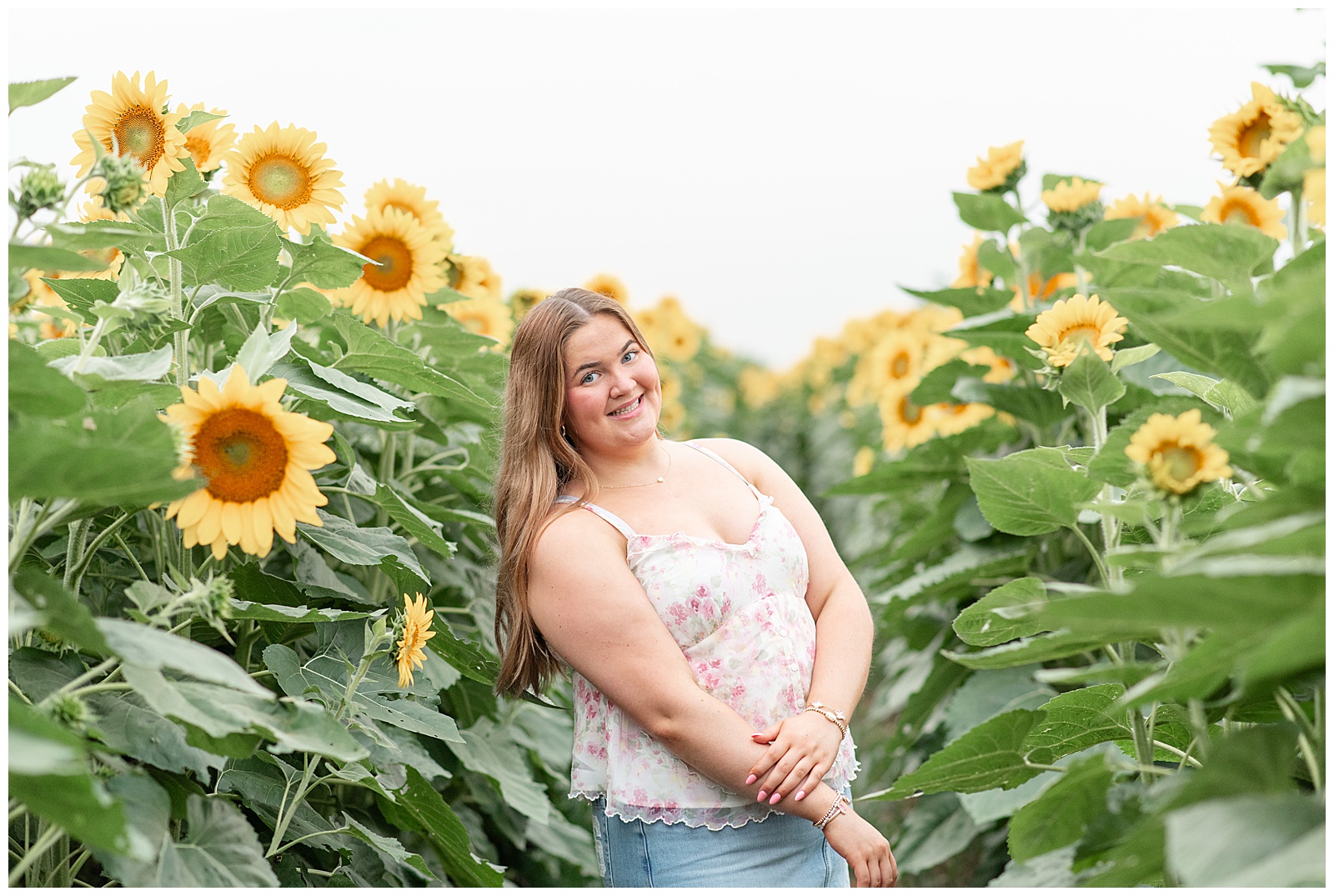 senior girl in white tank top and blue jean skirt by rows of sunflowers for this Cloudy Wildflower Field Senior Session