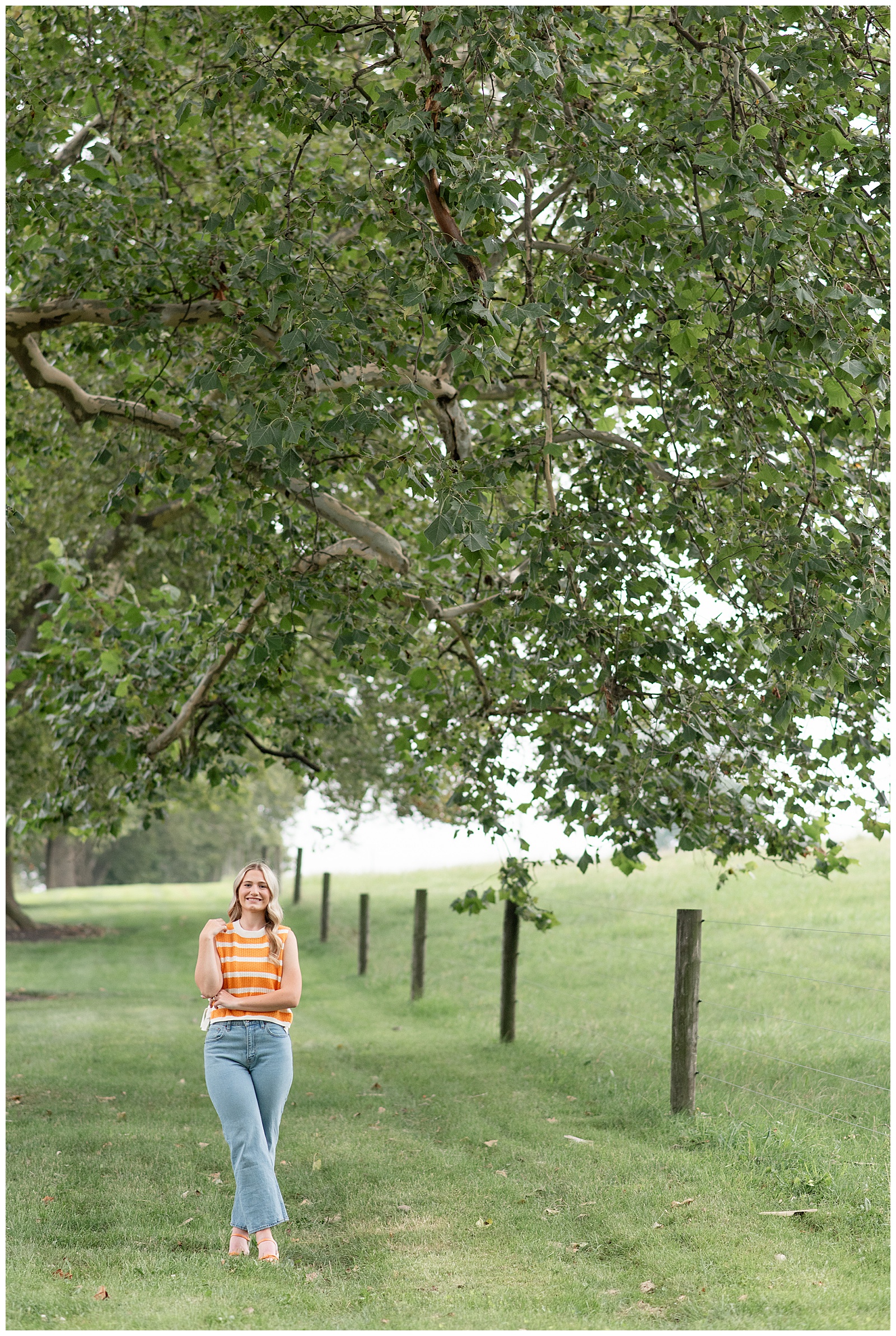 senior girl standing in grass with arms folded by fenced in pasture for this Masonic Village Summer Senior Session