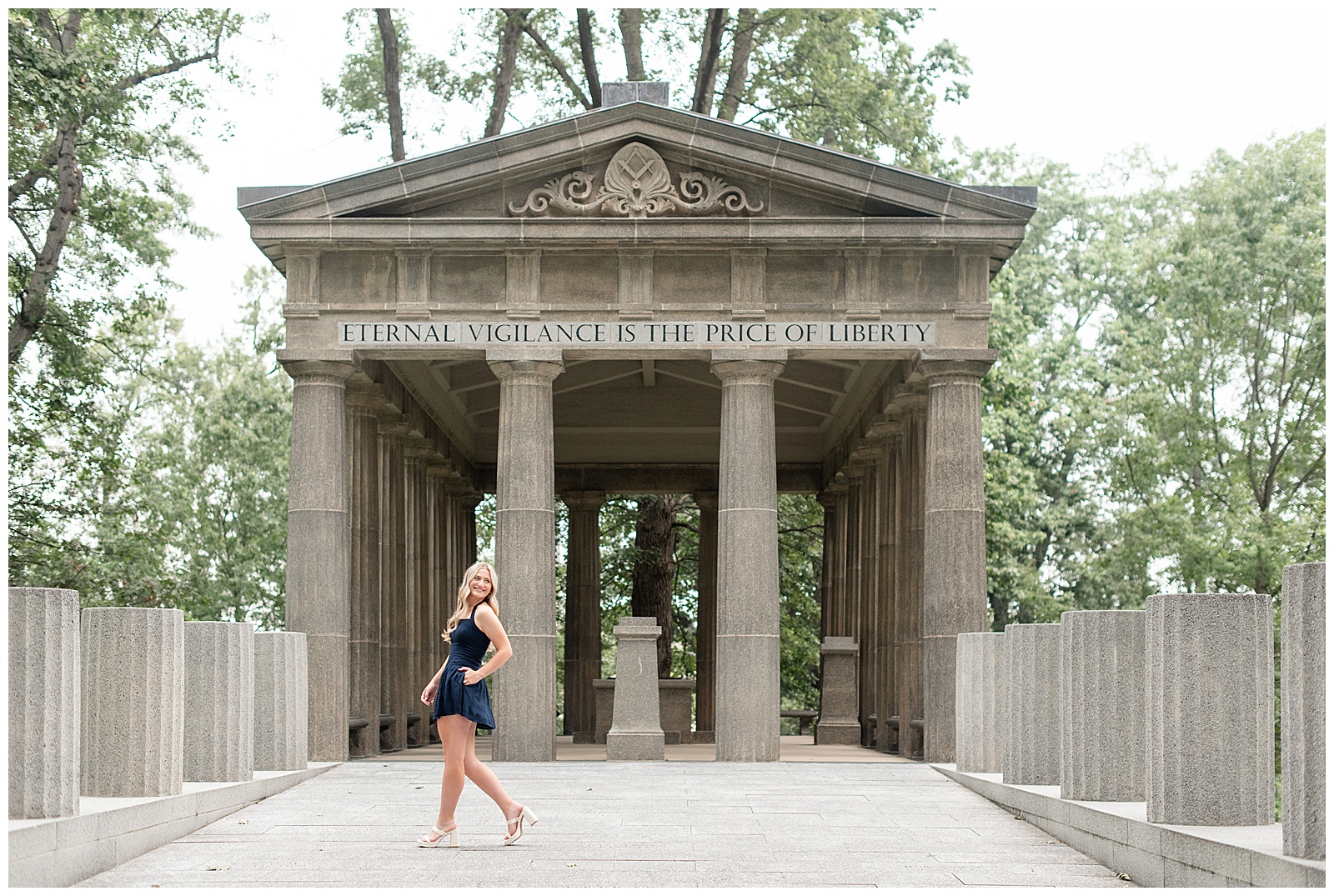 senior girl in navy blue dress walking and looking over left shoulder by large concrete structure at this Masonic Village Summer Senior Session