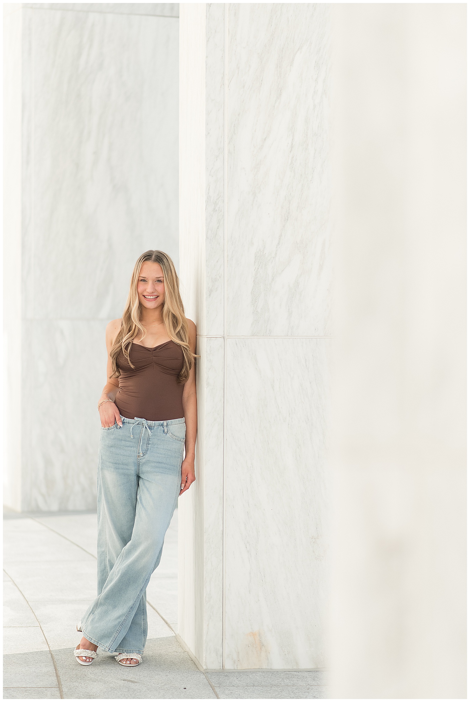 senior spokesmodel in brown tank top and blue jeans leaning again white wall for this Founders Hall Summer Senior Session