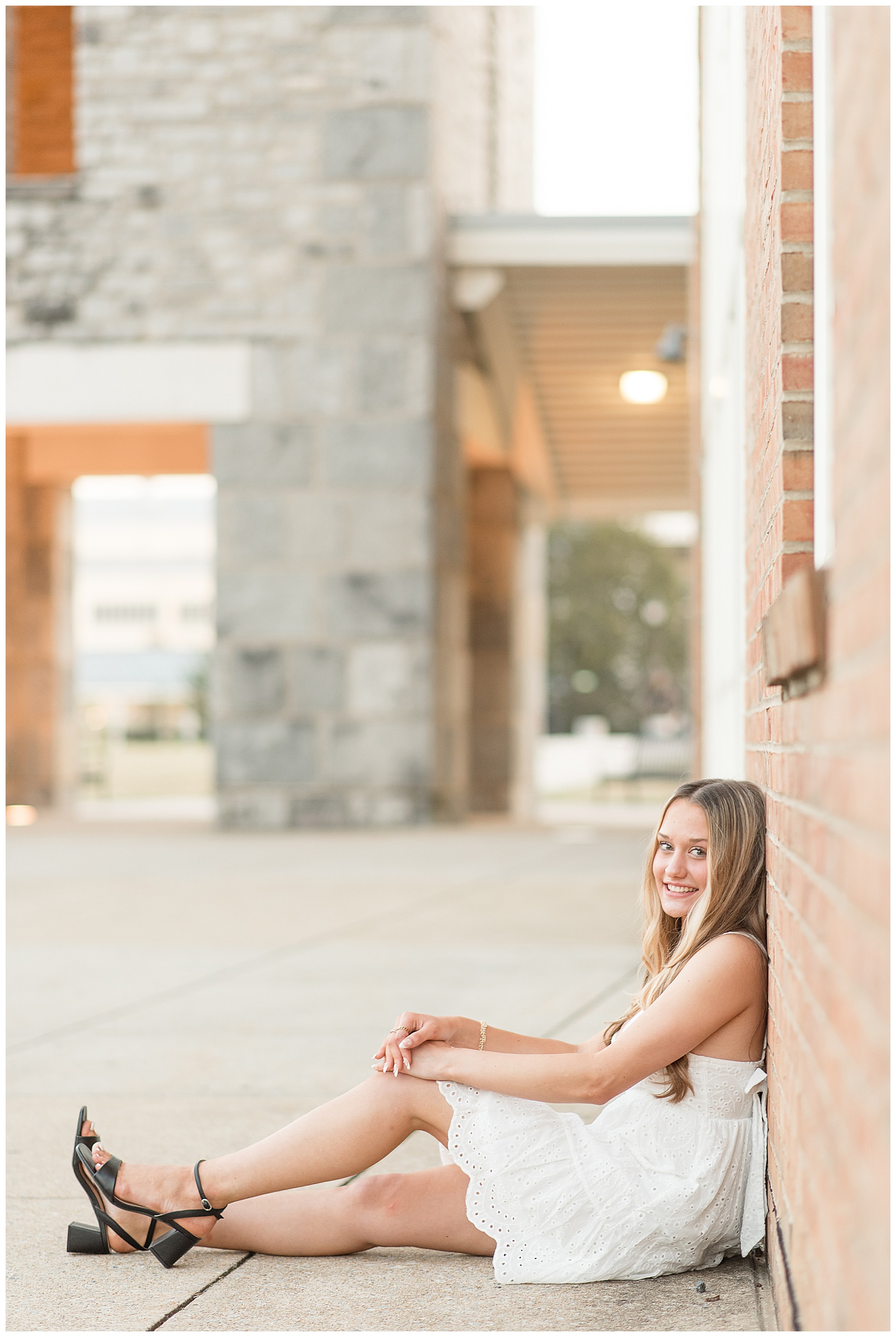 senior girl in white dress sitting with back against brick wall for this Founders Hall Summer Senior Session