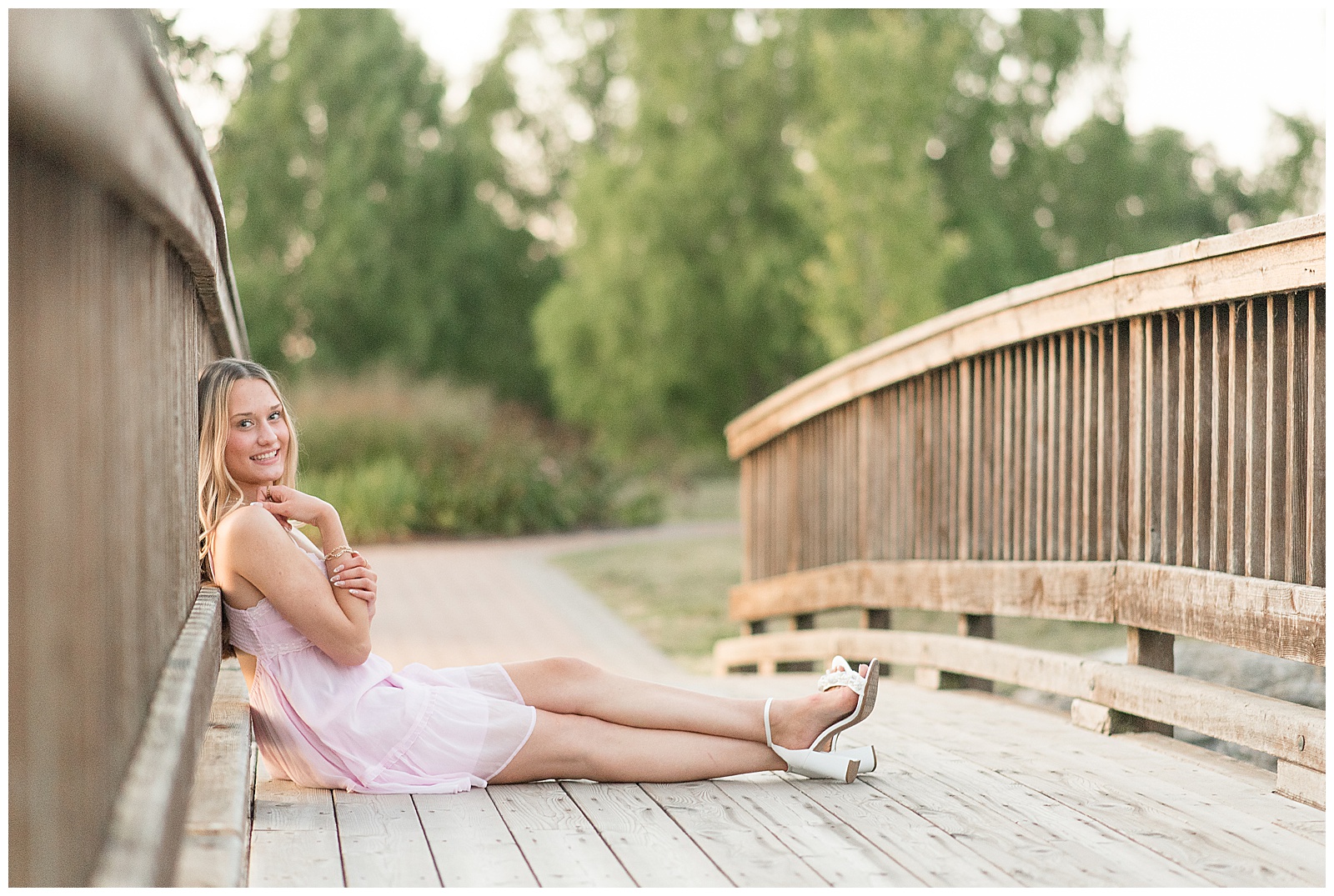 senior girl in white dress sitting on wooden bridge and leaning against rails for this Founders Hall Summer Senior Session